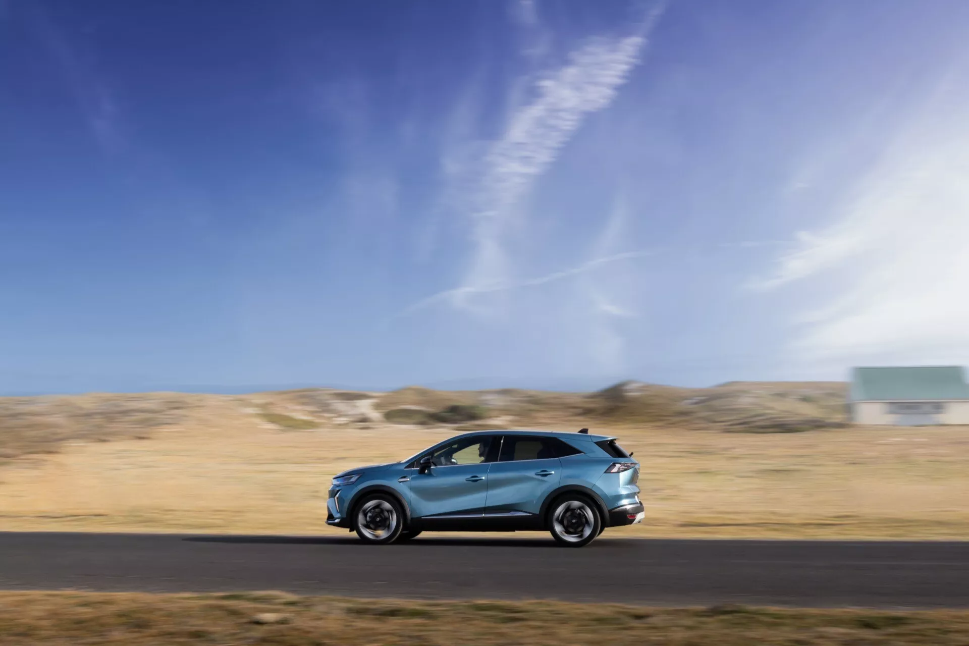 Dynamiczne ujęcie z profilu bocznego poruszającego się Renault Symbioz 2024 1.generacji, wykonane techniką panoramowania na rozmazanym tle, prezentujące aerodynamiczny design, alufelgi, lusterka boczne, zintegrowane klamki drzwi oraz wysokiej jakości lakier. Pojazd jest umieszczony w dolnej trzeciej części kadru na tle niebieskiego nieba i pustynnego krajobrazu, podkreślając prędkość, ruch i zdolność samochodu do działania w zróżnicowanych warunkach.