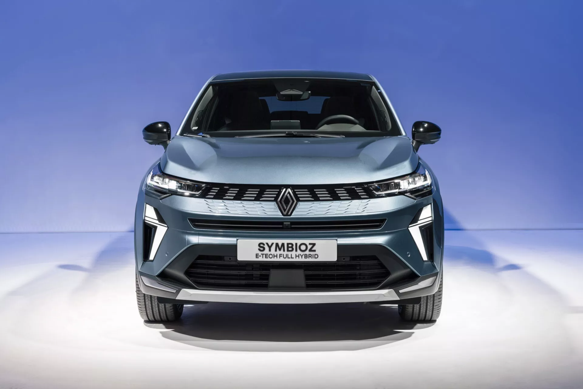 Zbliżenie przedniego widoku Renault Symbioz 2024 (1. generacja) uchwycone z lekkiego niskiego kąta. Obraz podkreśla elegancką przednią kratkę z stylizowanym emblematem 'R', kątowe reflektory LED i aerodynamicznie ukształtowaną maskę. Metaliczny jasnoniebieski lakier i błyszczące wykończenie samochodu są wyróżniane na tle czystego, przejściowego niebieskiego tła. Tekst 'SYMBIOZ' na obszarze tablicy rejestracyjnej sugeruje nowoczesny, prawdopodobnie futurystyczny design z funkcjami takimi jak oświetlenie LED, potencjalne systemy autonomiczne oraz zaawansowana łączność lub technologia napędu elektrycznego.