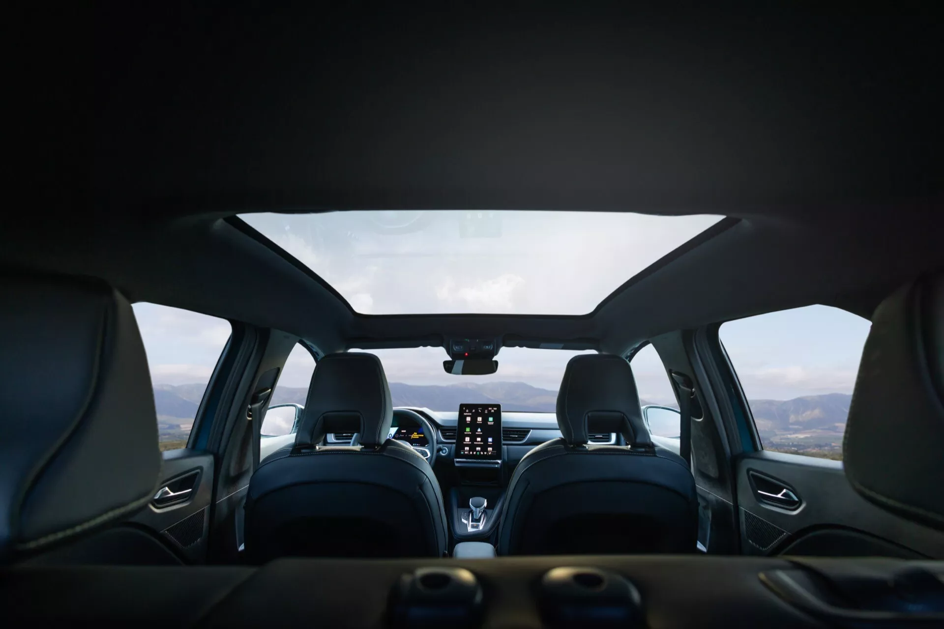 Wnętrze Renault Symbioz 2024 (1. generacja) widziane z perspektywy tylnego siedzenia, ukazujące nowoczesny design z skórzanymi siedzeniami, wyraźnymi szwami i wygodnie wyprofilowanymi przednimi fotelami z zagłówkami. Na centralnej desce znajduje się wyraźny pionowy ekran dotykowy systemu infotainment, pokrętła do sterowania klimatyzacją oraz cyfrowy wyświetlacz lub HUD na górze. Wielofunkcyjna kierownica jest częściowo widoczna. Widok przez przednią szybę ukazuje górski krajobraz pod pochmurnym niebem, sugerując, że pojazd jest zaparkowany w malowniczym miejscu.