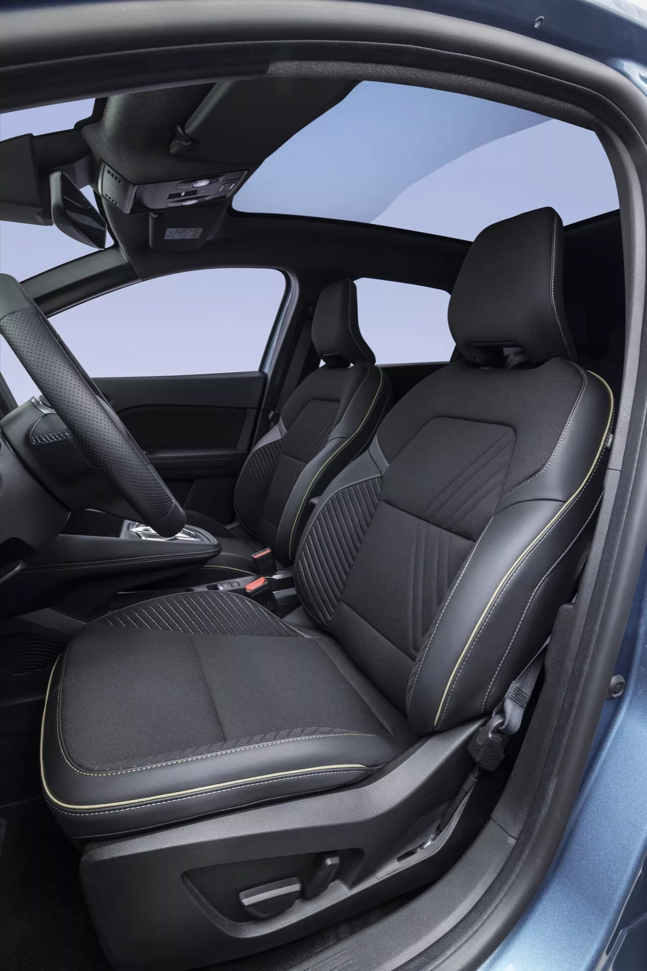 Obraz ukazuje wnętrze Renault Symbioz 2024 (1. generacja) z bocznej i lekko podniesionej perspektywy, koncentrując się na przednich siedzeniach kierowcy i pasażera. Siedzenia, tapicerowane ciemną skórą z pionowymi tłoczeniami, sugerują funkcje ogrzewania lub wentylacji dla zwiększenia komfortu. Wyraźne boczne wzmocnienia wskazują na projekt ukierunkowany na dynamiczną jazdę. Częściowo widoczny panel drzwiowy z ciemnym wykończeniem uzupełnia luksusową atmosferę i prawdopodobnie skrywa sterowniki okien i luster elektrycznych. Konsola środkowa jest wyposażona w dwa uchwyty na kubki, dźwignię zmiany biegów automatycznych i mały schowek. Widoczna część czystej, niezagraconej deski rozdzielczej zawiera centralny ekran infotainment, podkreślając nowoczesną technologię samochodową. Nieco widoczna wielofunkcyjna kierownica sugeruje dodatkowe funkcje technologiczne, takie jak sterowanie audio, polecenia głosowe i tempomat. Duży szklany panel w dachu sugeruje obecność szyberdachu lub dachu panoramicznego, co przyczynia się do przestronności kabiny. Ogólnie obraz przedstawia nowoczesne wnętrze z naciskiem na komfort, ergonomię i technologię, odpowiednie do kategorii wnętrz samochodowych, nowoczesnych technologii pojazdów i luksusowych cech samochodów.