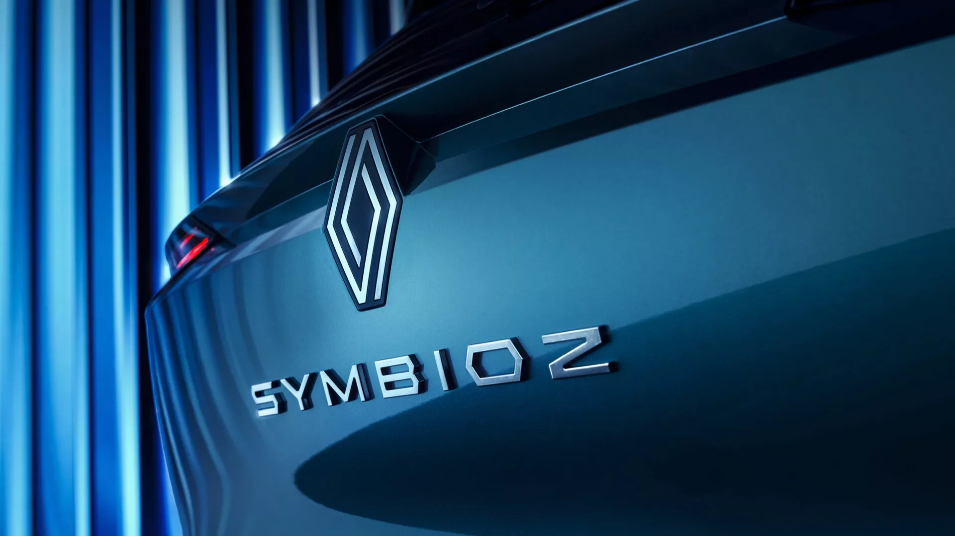 Zbliżony skośny widok tylnej części samochodu Renault Symbioz 2024 (1. generacja), z naciskiem na emblemat marki i nazwę modelu. Emblemat w kształcie diamentu i 'SYMBIOZ' wielkimi literami bezszeryfowym krojem są wyraźnie widoczne na pokrywie bagażnika, sugerując nacisk na innowacje i technologię. Miękki niebieski odcień rzutuje równoległe linie w tle, tworząc elegancką i nowoczesną atmosferę. Samochód wydaje się stać w miejscu, podkreślając jego aerodynamiczne linie projektowe.