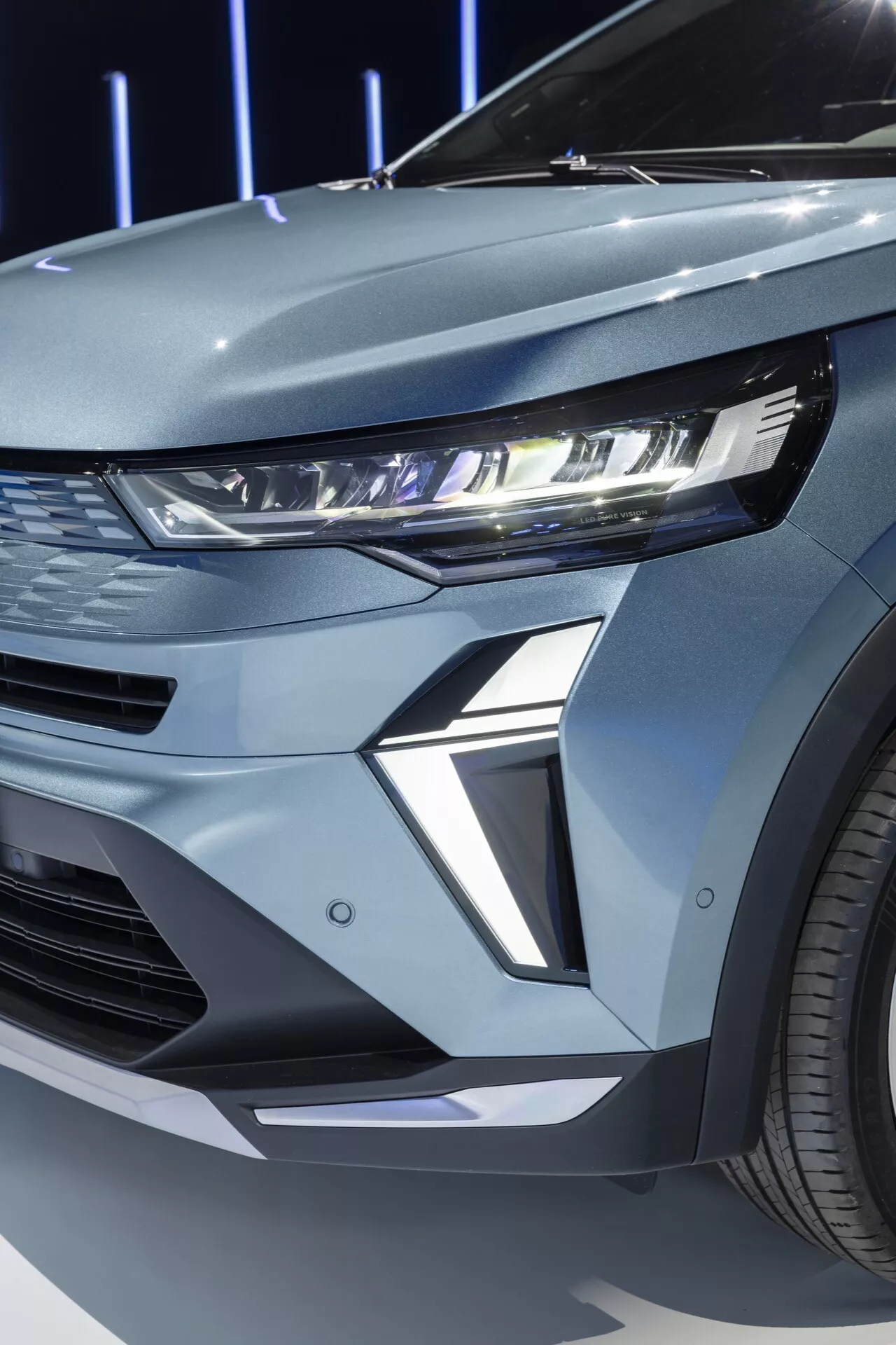Zbliżenie przedniego lewego kwadrantu Renault Symbioz 2024 pierwszej generacji. Na zdjęciu widoczny jest nowoczesny reflektor LED pojazdu z podświetloną sygnaturą światła do jazdy dziennej (DRL). Wąska obudowa reflektora rozciąga się w kierunku błotnika, co jest przykładem współczesnego designu samochodowego. Rzeźbiona sekcja pod reflektorem dodaje pojazdowi futurystyczny i agresywny wygląd. Widoczny segment przedniego grilla sugeruje poziome wloty powietrza lub panel dekoracyjny, zgodny ze sportową estetyką. Metaliczne wykończenie pojazdu i widoczne wieloszprychowe felgi wzmacniają jego luksusowy wygląd.