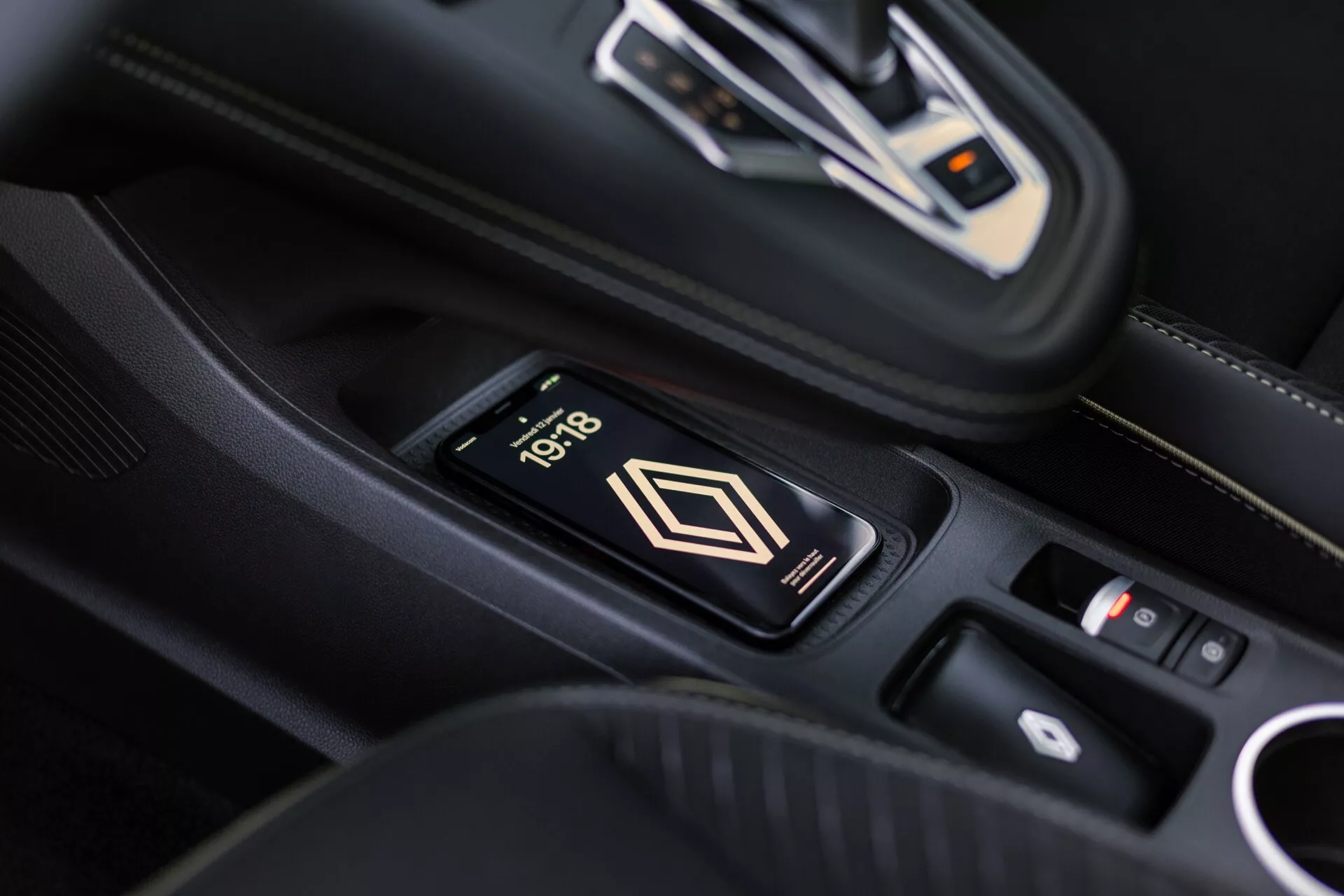 Wnętrze Renault Symbioz 2024, widok na konsolę centralną z bezprzewodową ładowarką do smartfonów, na ekranie wyświetlany jest czas 10:18. W tle widoczna automatyczna dźwignia zmiany biegów z metalicznymi akcentami i panel sterowania. Wnętrze premium w czarnej skórze z białymi szwami.