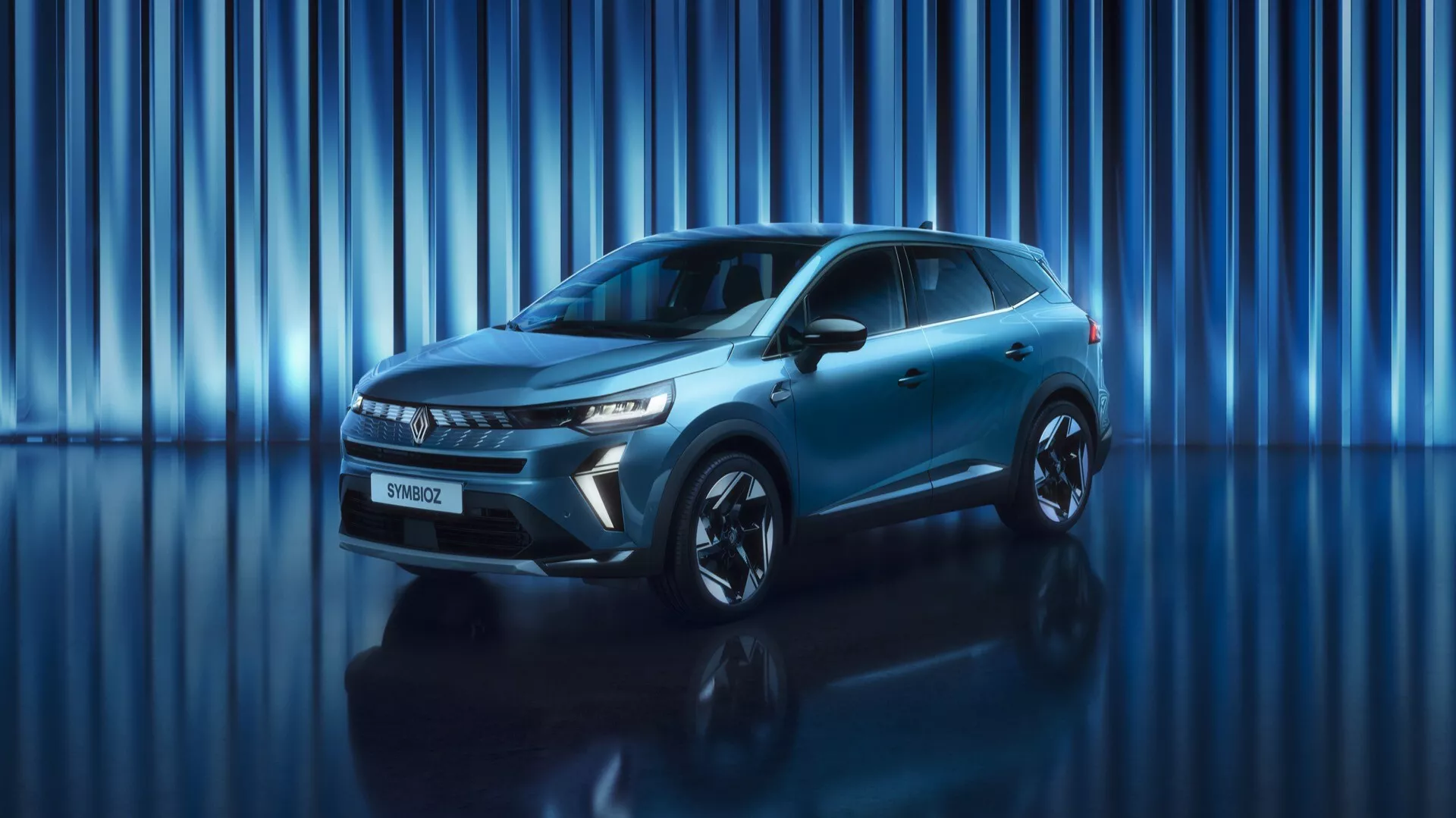 Obraz przedstawia Renault Symbioz 2024 (1. generacji) SUV ustawiony w studio z dynamicznym niebieskim tłem z pionowymi pasami świetlnymi. Kadr z niskiego do średniego kąta ukazuje potężny przód i smukły profil boczny pojazdu. Cały samochód jest w kadrze, podkreślając jego obecność i nowoczesny design. Widoczne są ostre, kątowe reflektory LED oraz wyraźny grill, a także gładki przedni zderzak, który prawdopodobnie zawiera czujniki bezpieczeństwa. Profil boczny ujawnia aerodynamiczne linie, płaskie klamki drzwi, smukłe lusterka boczne i duże felgi aluminiowe. Metaliczna powłoka i sportowa sylwetka samochodu są podkreślone przez oświetlenie studyjne oraz odbijającą powierzchnię pod nim.
