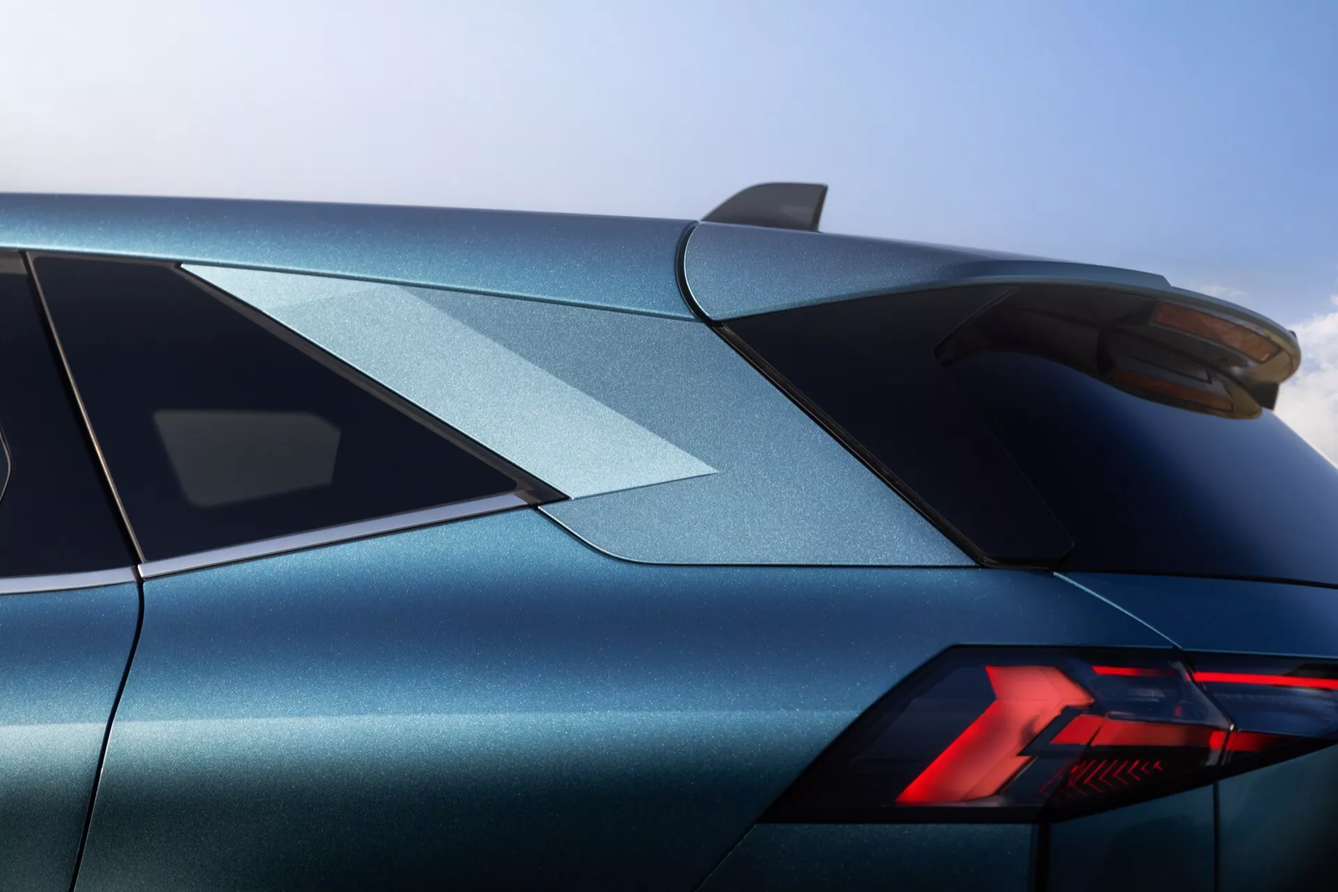 Zbliżony widok zza trzech czwartych tylnej części Renault Symbioz 2024, uwydatniający jego górną tylną część. Obraz przedstawia charakterystycznie stylizowane tylne światło z nowoczesnymi elementami LED w układzie 'Y'. Elegancka karoseria, wykończona połyskującą metaliczną farbą w kolorze niebiesko-szarym, ma ostre, wyraźnie zarysowane linie. Mocno pochylona tylna szyba i zamontowany tylny spojler podkreślają sportową estetykę samochodu. Na dachu widoczna jest antena w kształcie płetwy rekina, co wskazuje na nowoczesne elementy designu. Wysoka jakość wykonania i precyzyjne detale są widoczne z tego kąta, sugerując luksusowy lub sportowy pojazd.