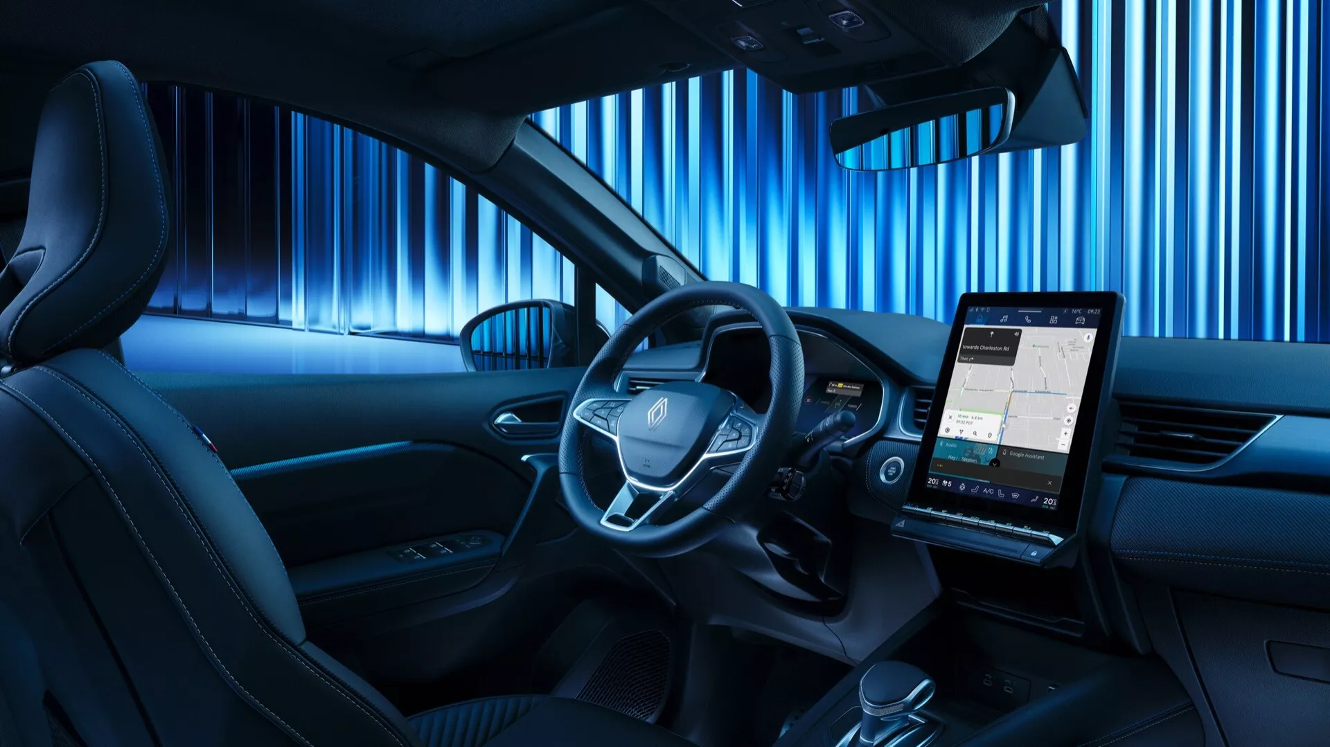 Widok z tylnego siedzenia na wnętrze Renault Symbioz 2024, z centralnie umieszczoną kierownicą z zintegrowanymi przyciskami sterującymi, cyfrowym panelem zegarów i wyraźnym pionowym ekranem infotainment wyświetlającym mapę. Kabina cechuje się niebieskim oświetleniem ambientowym, sugerującym nowoczesny i technologiczny design, minimalną ilością fizycznych przycisków na centralnej konsoli oraz częścią panelu drzwiowego z wbudowaną kratką głośnika, co wskazuje na wysokiej jakości system dźwiękowy.