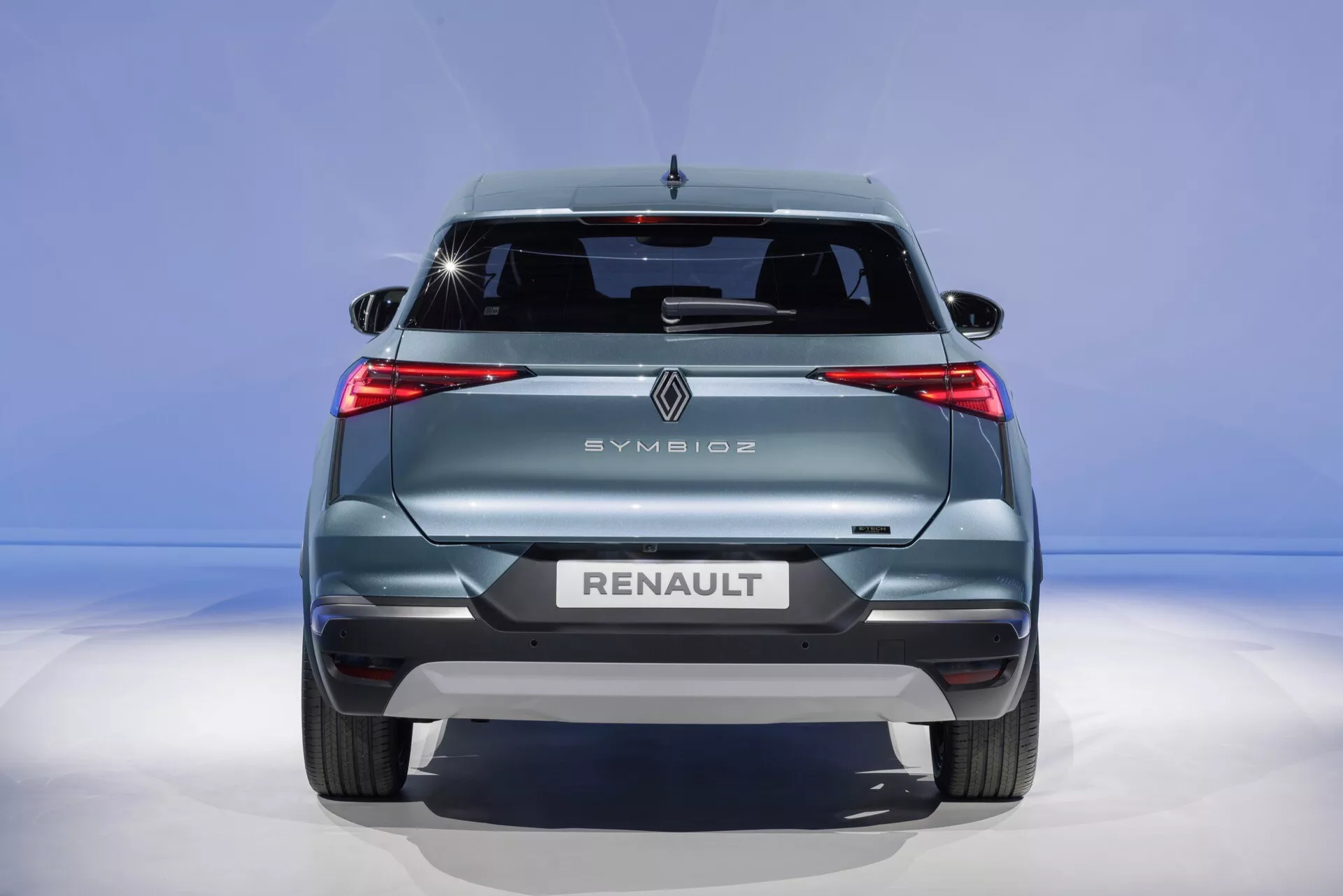 Widok z tyłu Renault Symbioz 2024, prezentujący nowoczesne elementy designu, w tym charakterystyczne tylne światła LED, wyraźnie widoczne logo marki, wypisany wielkimi literami model i markę, tylna wycieraczka, wysoko zamontowane światło stop oraz czujniki zderzaka tylnego na niebieskim gradientowym tle.