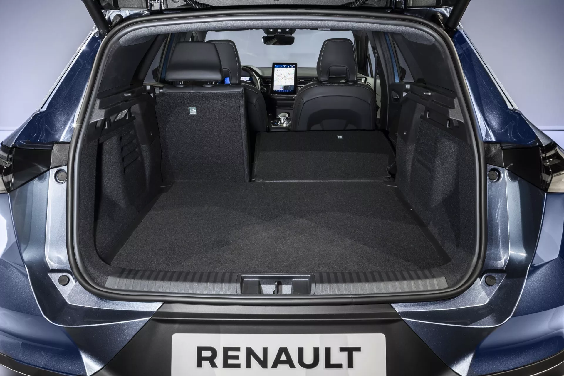 Widok na przestrzeń bagażową Renault Symbioz 2024 z wysokiego kąta za otwartą tylną klapą, pokazujący czysty, uporządkowany bagażnik z pionowymi siedzeniami, tylnym zderzakiem z oznaczeniem Renault, trwałą wykładziną, gniazdem zasilania 12 V po lewej stronie i mocowaniem do zabezpieczania przedmiotów po prawej. Widoczny jest wnętrze kabiny oraz ekran konsoli centralnej, z równomiernym oświetleniem studyjnym podkreślającym cechy.