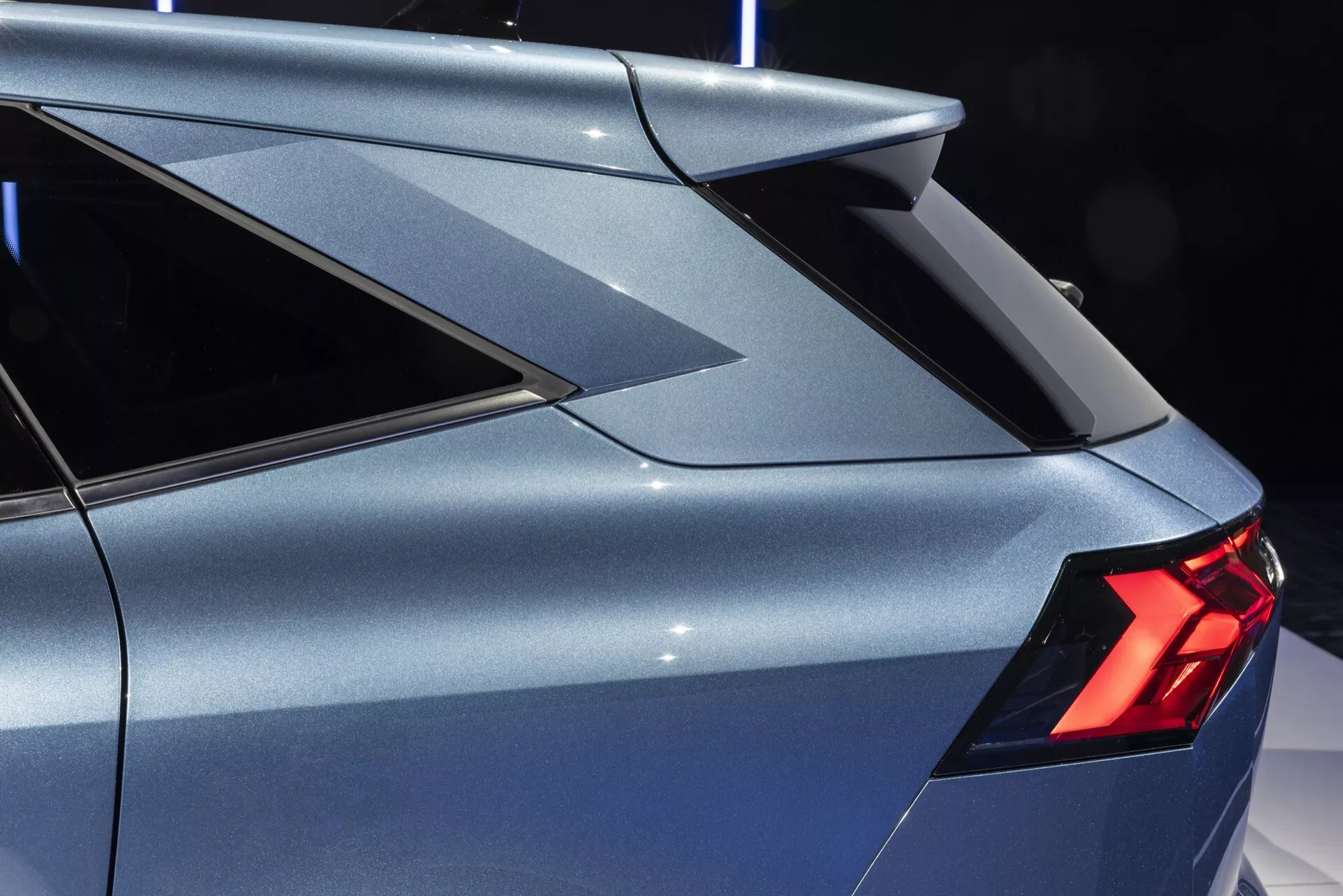 Zbliżenie na tylną sekcję Renault Symbioz 2024 (1. generacja). Obraz ukazuje pojazd z lekko podwyższonego bocznego kąta, podkreślając ostry, kanciasty słupek C z zintegrowanym, sportowym spojlerem tylnym. Widoczne są wąskie, wyraźne tylne światła LED, a także część tylnego drzwi pasażera oraz subtelnie zakrzywiona, aerodynamiczna karoseria. Metaliczna powłoka lakiernicza odbija oświetlenie otoczenia, co sugeruje jasne, profesjonalnie oświetlone miejsce.