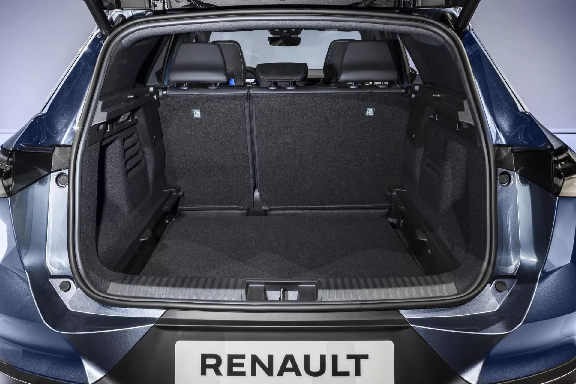 Widok wnętrza przestrzeni ładunkowej Renault Symbioz 2024 od strony tylnego klapy, pokazujący symetryczną przestrzeń bagażnika z czarnym dywanikiem, plastikowymi wykończeniami oraz złożonymi tylnymi siedzeniami dla zwiększonej pojemności. Widoczna marka obejmuje 'RENAULT' poniżej klapy.