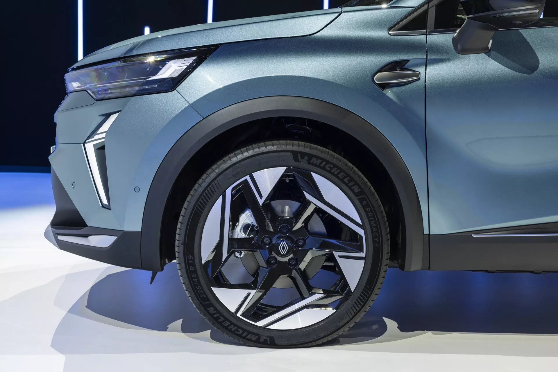 Zbliżenie przedniej sekcji Renault Symbioz 2024 (1. generacja), podkreślające nowoczesne czarne aluminiowe koło z wieloszprychowym wzorem, niskoprofilową oponę z widocznymi literami na ścianie bocznej oraz wyraźne, muskularne linie błotnika. Obraz uchwycił także niewielką część drzwi z charakterystyczną linią oraz kanciasty, elegancki design świateł na przedniej fasadzie. Lakier samochodu wydaje się matowy lub satynowy, co może sugerować model premium lub edycję specjalną.