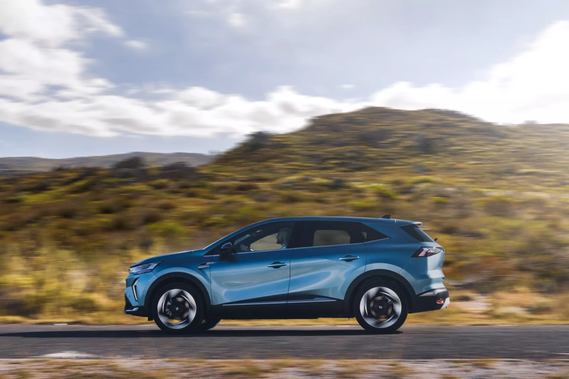 Jasnoniebieski Renault Symbioz 2024 (1. generacja) w ruchu, ukazujący swoje eleganckie linie aerodynamiczne i nowoczesny design z profilu. Samochód, umieszczony w lewej trzeciej części obrazu, ma rozmyte tło i drogę, co wskazuje na prędkość. Widoczne cechy to LED-owe światła do jazdy dziennej, zintegrowane kierunkowskazy w lusterkach bocznych, panoramiczny dach, wyraziste felgi oraz opony o niskim profilu. Zdjęcie wykonane za pomocą techniki panningu, która podkreśla dynamiczny ruch samochodu.