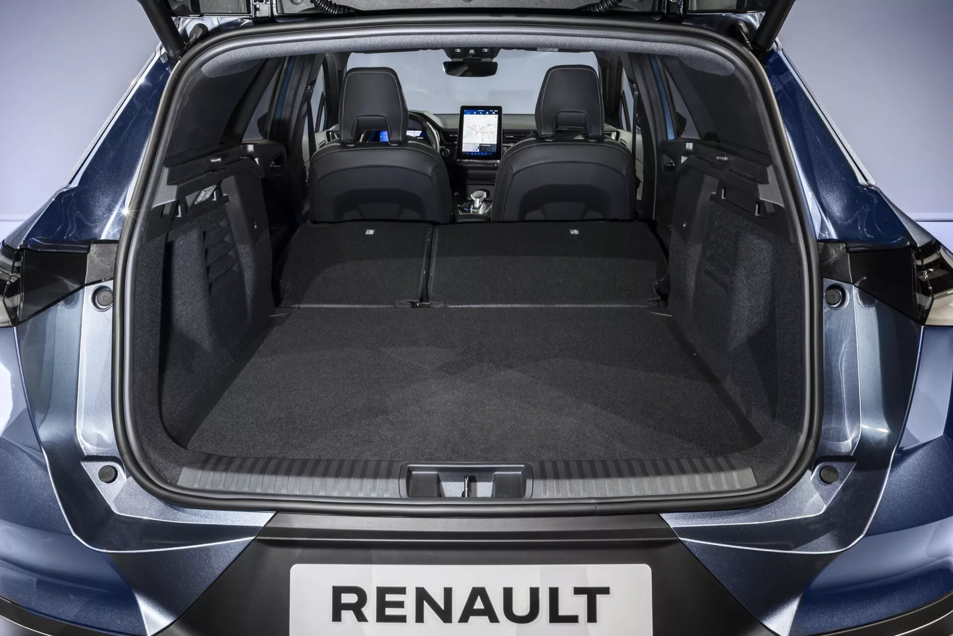 Widok od tyłu przez otwarty bagażnik do przestrzeni bagażowej Renault Symbioz 2024. Przestronny bagażnik ma płaską podłogę wyłożoną szarym materiałem przypominającym dywan, zaprojektowaną bez przeszkód dla maksymalnej pojemności. Dwa zatrzaski po obu stronach sugerują możliwość złożenia tylnych siedzeń w celu uzyskania większej przestrzeni. Czarne skórzane tylne siedzenia z zagłówkami oraz ledwo widoczny nowoczesny system informacyjno-rozrywkowy w przedniej kabinie sugerują wnętrze wyposażone w technologię.