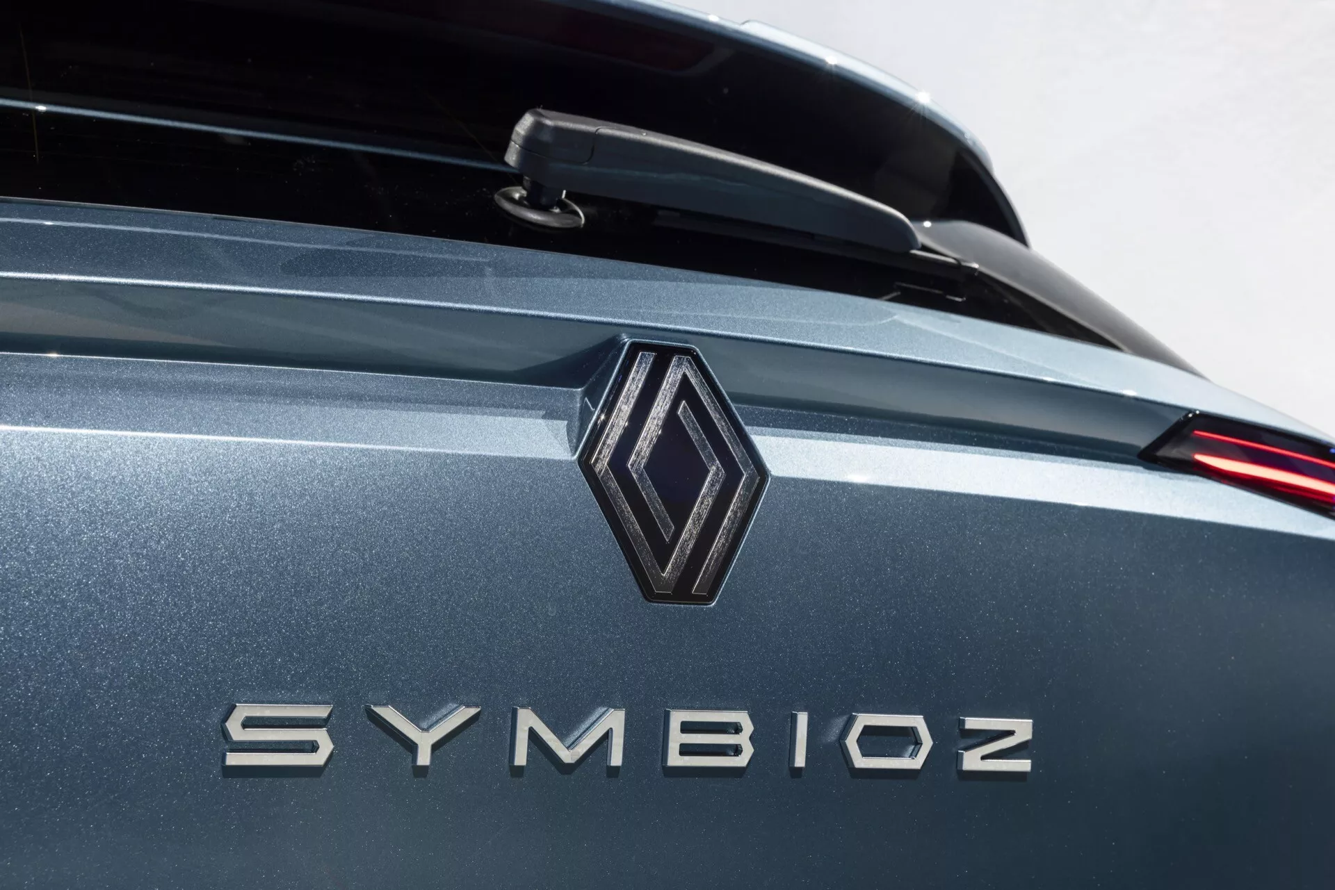 Profesjonalne zbliżenie tylnej części samochodu skupiające się na detalach marki z emblematem w kształcie diamentu i nazwą modelu 'SYMBIOZ' w metalicznych literach. Zdjęcie wykorzystuje płytką głębię ostrości, podkreślając emblemat i litery, podczas gdy tło łagodnie się rozmywa. Dobrze oświetlone otoczenie sugeruje wewnętrzny salon lub studio. Kolor pojazdu to metaliczne wykończenie w odcieniach szarości lub niebieskiego, z możliwością obecności tylnego spojlera i nowoczesnym designem tylnych świateł LED.