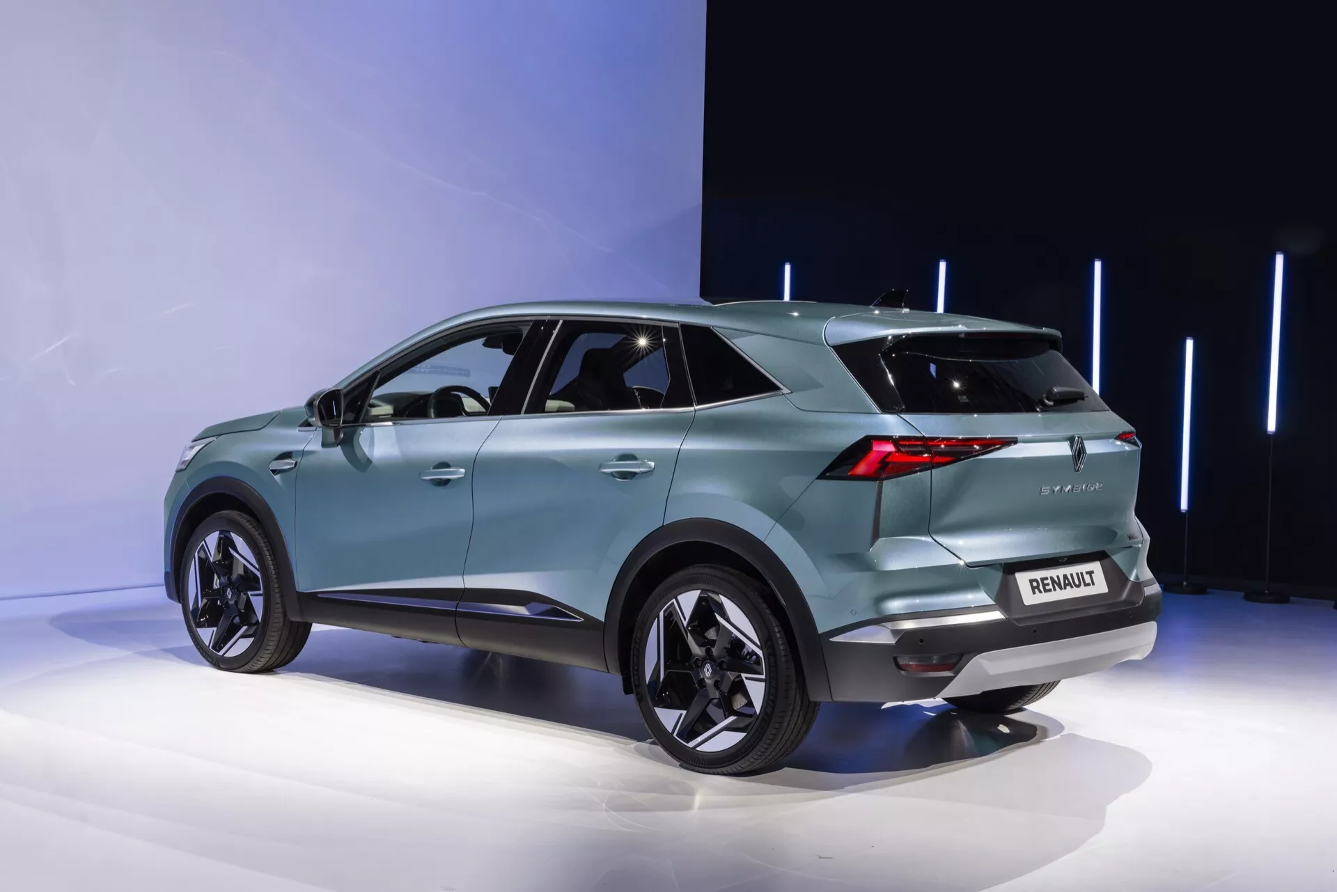 Widok tylno-boczny Renault Symbioz 2024. Turkusowy SUV, ustawiony na platformie wystawowej, prezentuje nowoczesny, aerodynamiczny design z pochylonym dachem, wyraźnymi nadkolami mieszczącymi duże aluminiowe felgi oraz przyciemnianymi szybami tylnymi. Wyróżniające się tylne światła LED, tylni spoiler, widoczne logo i oznaczenie modelu, czujniki parkowania oraz możliwa tylna kamera są widoczne. Elegancki tylny zderzak zawiera również elementy oświetleniowe, a antena typu płetwa rekina sugeruje zaawansowane technologie. Zintegrowane kierunkowskazy w lusterkach zewnętrznych i wpuszczone klamki drzwi wskazują na bezkluczykowy dostęp. Minimalistyczne tło podkreśla detale pojazdu.
