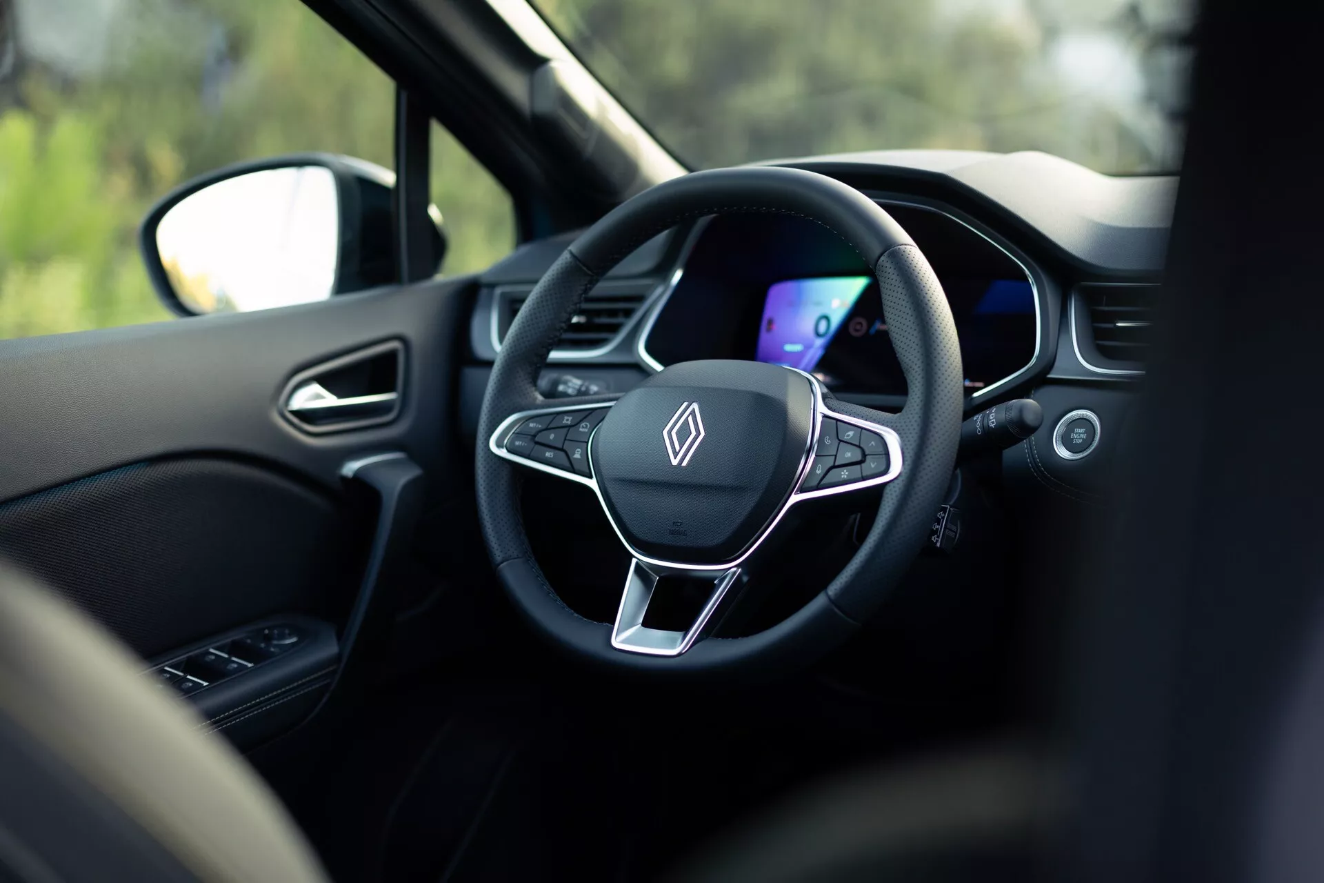 Widok wnętrza Renault Symbioz 2024 z tylnego siedzenia, skupiający się na nowoczesnej, trójramiennej kierownicy i cyfrowym zestawie wskaźników. Kierownica posiada płaskie dno, metaliczny akcent i zintegrowane przyciski sterujące, otoczone ciemną kolorystyką. Płytka głębia ostrości podkreśla te elementy, z rozmytym otoczeniem, w tym zagłówkiem i widocznym za oknem krajobrazem, sugerującym, że samochód jest zatrzymany.