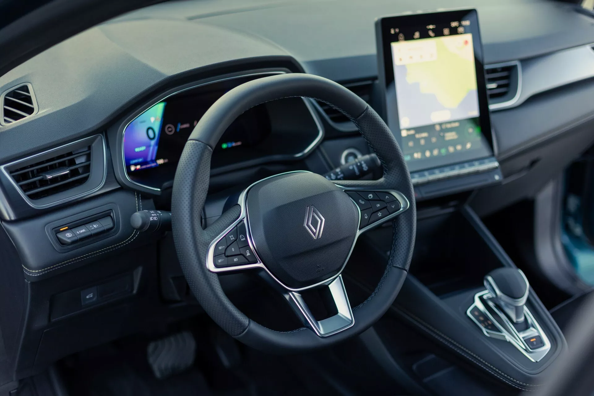 Zdjęcie wnętrza Renault Symbioz 2024 (1. generacja) z perspektywy kierowcy, skupiające się na kierownicy i desce rozdzielczej. Zdjęcie przedstawia widok z lotu ptaka pod kątem 30 stopni w dół, pokazujące trójramienną kierownicę z wielofunkcyjnymi przyciskami i ogólnym emblematem, w pełni cyfrowy zestaw wskaźników i duży ekran dotykowy systemu informacyjno-rozrywkowego wyświetlający mapę. Konsola środkowa ma nowoczesny selektor biegów o minimalistycznym designie. Wnętrze ma zaawansowaną technologicznie konstrukcję elektrycznego pojazdu z oświetleniem ambientowym i nowoczesną estetyką.