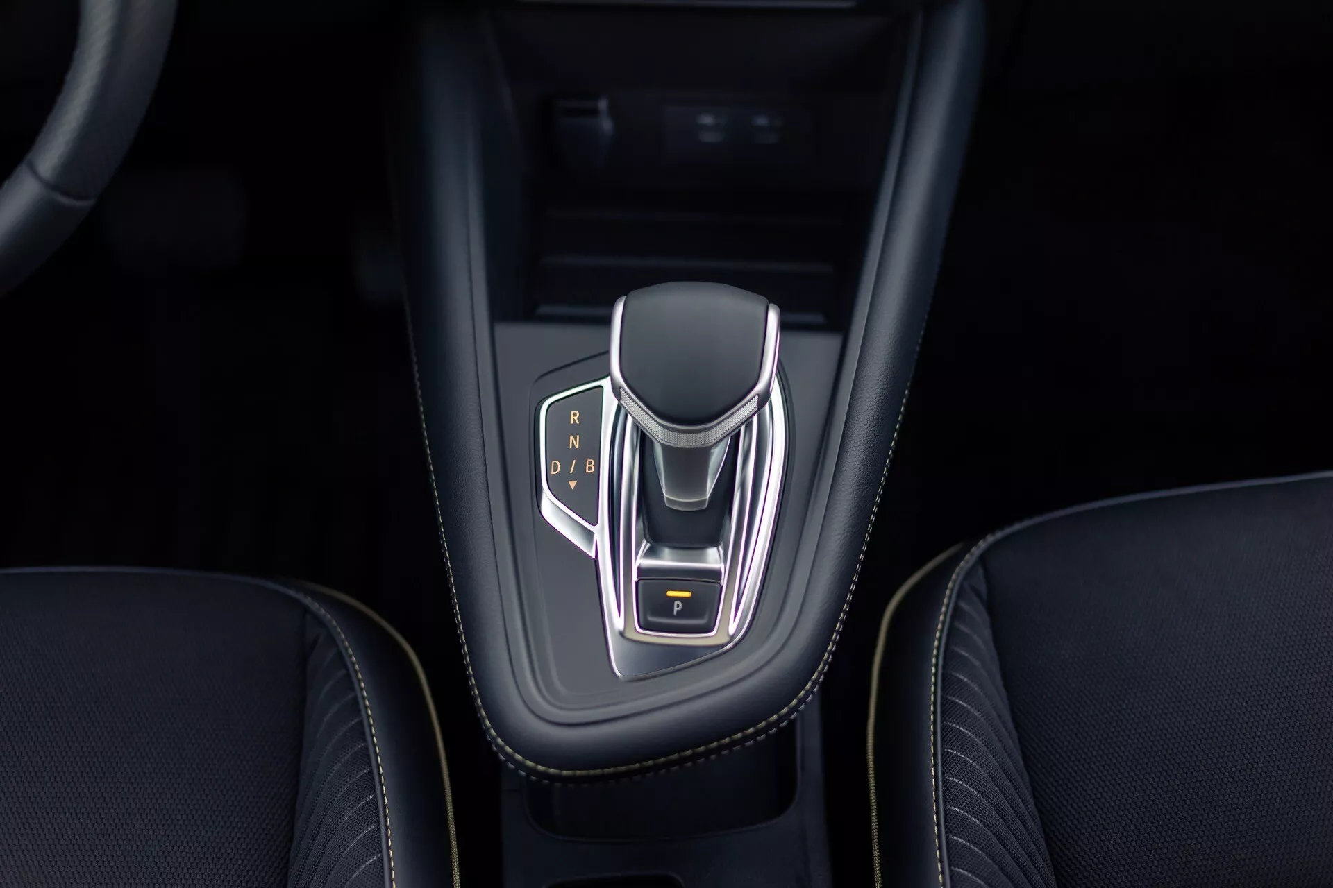 Widok z góry na konsolę centralną Renault Symbioz 2024, skoncentrowany na elektronicznym wybieraku biegów z chromowanym wykończeniem i podświetlanymi wskaźnikami biegów dla trybów Park, Reverse, Neutral i Drive. Wybierak ma boki pokryte skórą, żółty podświetlany przycisk parkowania i jest osadzony w błyszczącej czarnej obudowie z misternym szwem na otaczającym skórzanym wnętrzu.