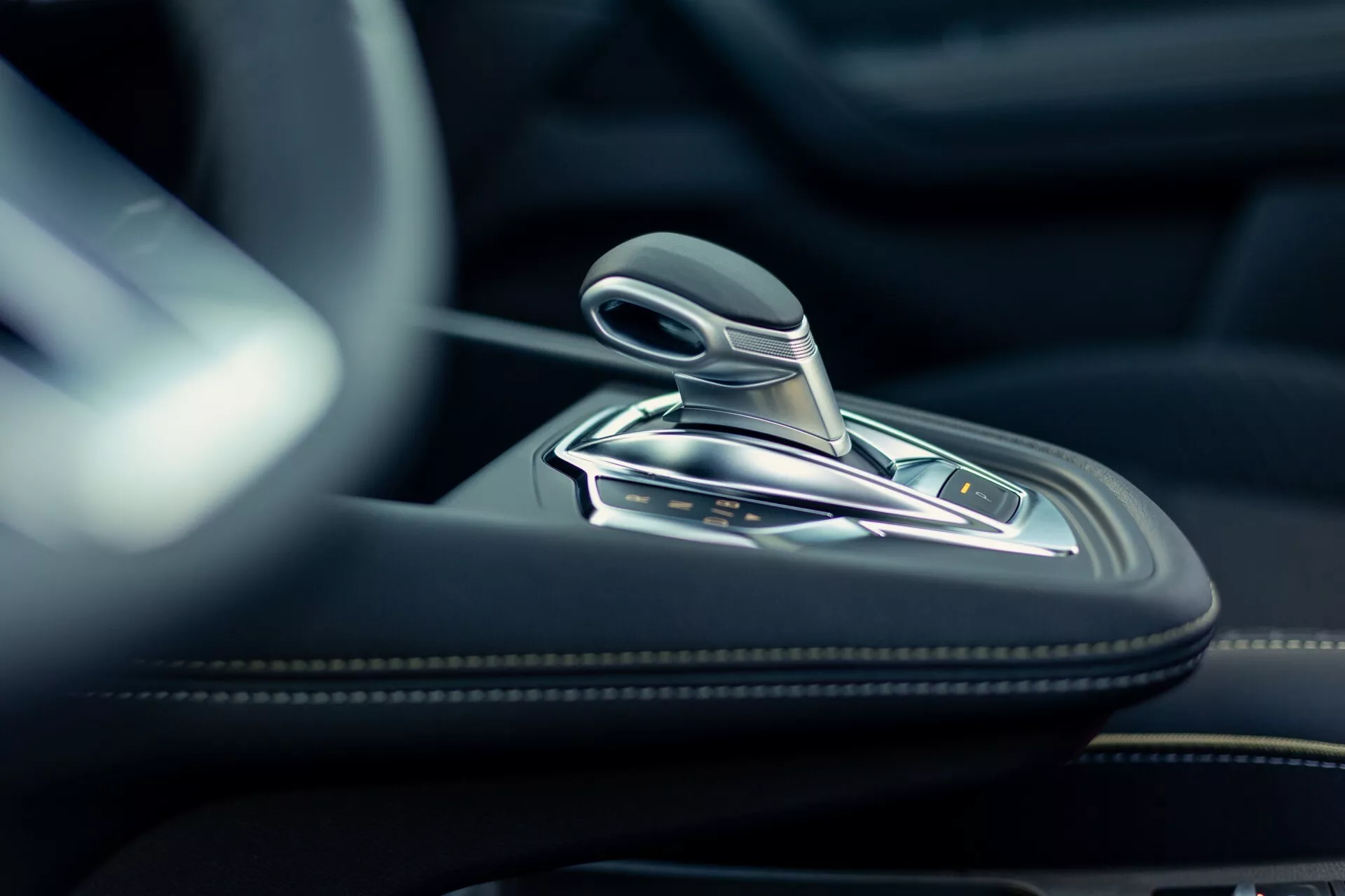 Zbliżenie wnętrza Renault Symbioz 2024 pokazujące nowoczesną dźwignię zmiany biegów z metalowymi akcentami i prawdopodobnie skórzanym wykończeniem. Zdjęcie wysokiej rozdzielczości wykonane z niskiego kąta koncentruje się na selektorze automatycznej skrzyni biegów i otaczającej konsoli centralnej, wykonanej z wysokiej jakości materiałów, takich jak skóra, szczotkowany metal lub chrom. Na konsoli widoczne są różne przyciski prawdopodobnie do trybów jazdy, hamulca ręcznego, funkcji auto-hold i kontroli trakcji. Miękkie, naturalne oświetlenie podkreśla luksusowe i precyzyjne wykonanie wnętrza samochodu.