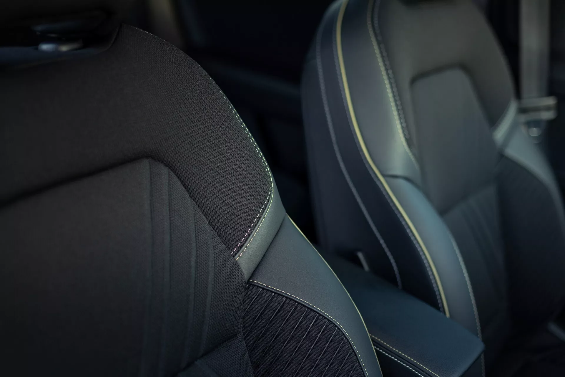 Zbliżenie przedniego fotela pasażera w Renault Symbioz 2024 (1. generacja). Fotel ma środkowy panel wykonany z tkaniny teksturowanej lub syntetycznego materiału zwiększającego oddychalność, otoczony gładkim, prawdopodobnie syntetycznym materiałem przypominającym skórę z kontrastującym ozdobnym szwem, co podkreśla jego luksusowy lub sportowy wygląd. Płytka głębia ostrości skupia się na fotelu, rozmywając tło, aby podkreślić jakość materiału i kunszt wykonania.