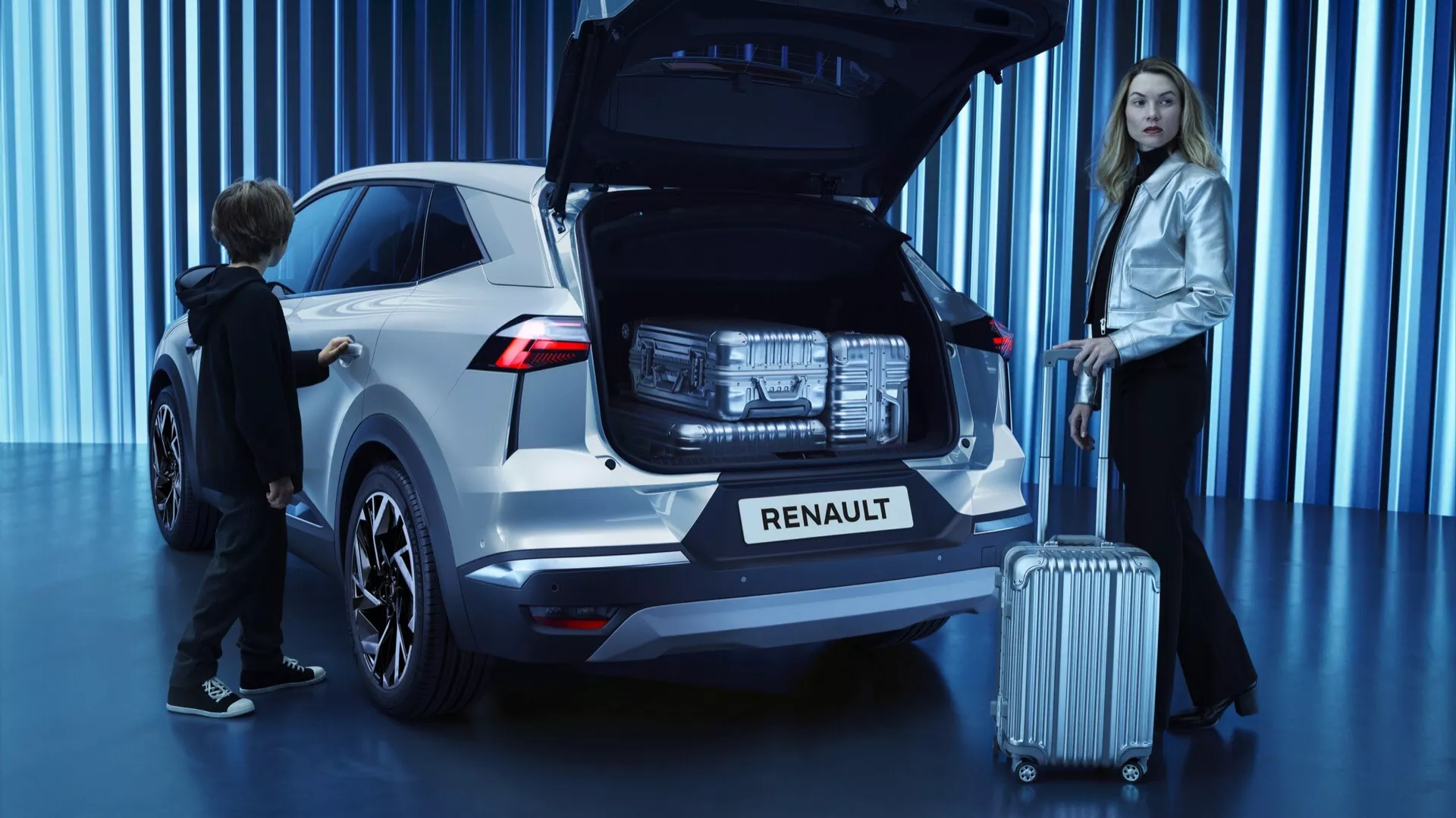 Widok trzech czwartych z tyłu białego SUV-a Renault Symbioz 2024 w salonie samochodowym, z otwartą klapą tylną ukazującą przestronny bagażnik, nowoczesne tylne światła LED, logo Renault pod tylną szybą, czujniki wspomagające parkowanie na zderzaku, wieloszprychowe felgi aluminiowe, system automatycznego otwierania przyciskiem oraz dwie osoby: kobieta z walizką sugerująca podróż i dziecko wchodzące w interakcję z bagażnikiem, podkreślające rodzinny charakter pojazdu.