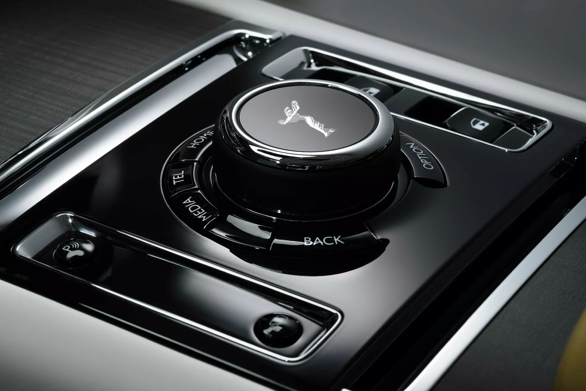 Zbliżony widok konsoli środkowej Rolls-Royce Spectre 2023 pierwszej generacji, pokazujący pokrętło sterowania systemem infotainment z powierzchnią dotykową, otoczone przyciskami z wyraźnie oznaczonymi funkcjami na panelu w wysokim połysku czarnym, osadzone w szczotkowanym metalu dla luksusowego designu wnętrza