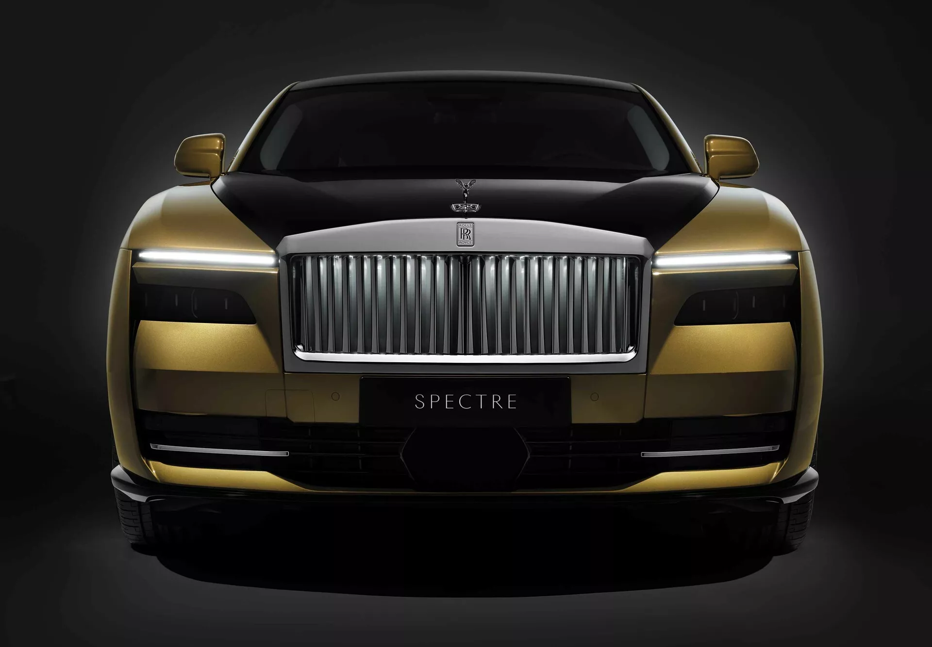 Rolls-Royce Spectre pierwszej generacji 2023, prezentujący dwukolorowy schemat malowania, zaawansowaną technologię reflektorów LED i aerodynamiczny design z widoczną nazwą modelu 'SPECTRE', uchwycony z niskiego, czołowego kąta na ciemnym tle.