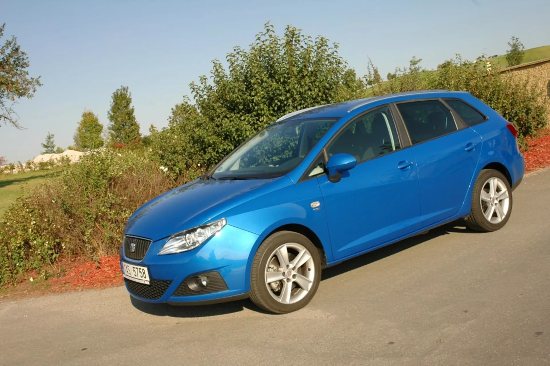 Jasnoniebieskie hatckback Seat Ibiza 2012 zaparkowane na asfalcie przeciwko zieleni, prezentujące swoją aerodynamiczną przednią kratę, reflektory zwrócone do tyłu i boczny profil z wieloramienne felgi aluminiowe pod naturalnym światłem dziennym