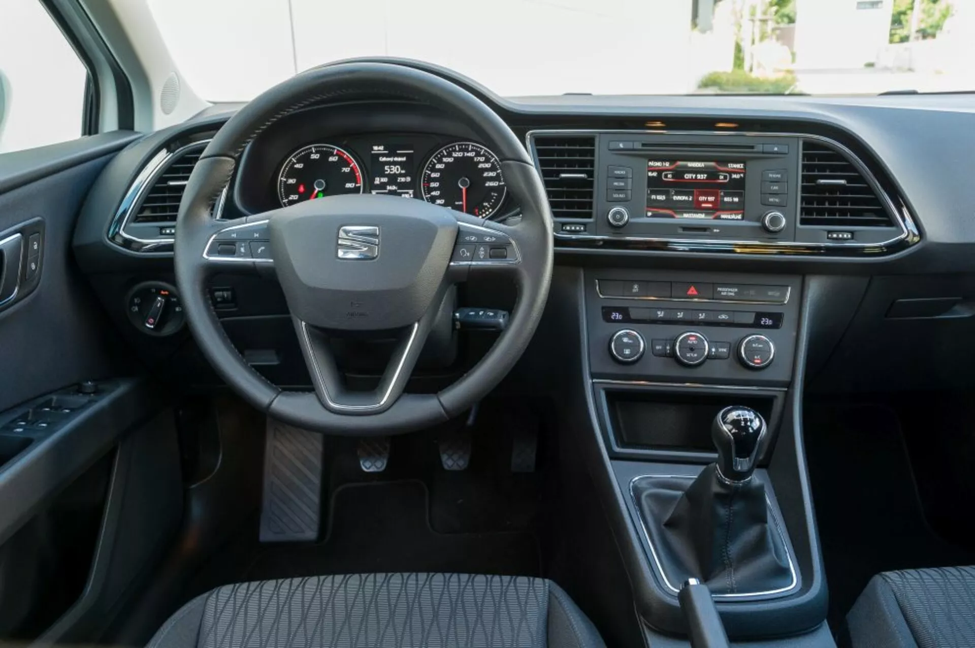 Widok wnętrza Seat Leon SC z 2016 roku z perspektywy przedniego siedzenia pasażera, prezentujący kierownicę z logo producenta i zamontowane kontrolki, cyfrowy kokpit, system infotainment, kontrolki klimatyzacji, manualną dźwignię zmiany biegów oraz drzwi kierowcy z opcjami elektrycznymi.