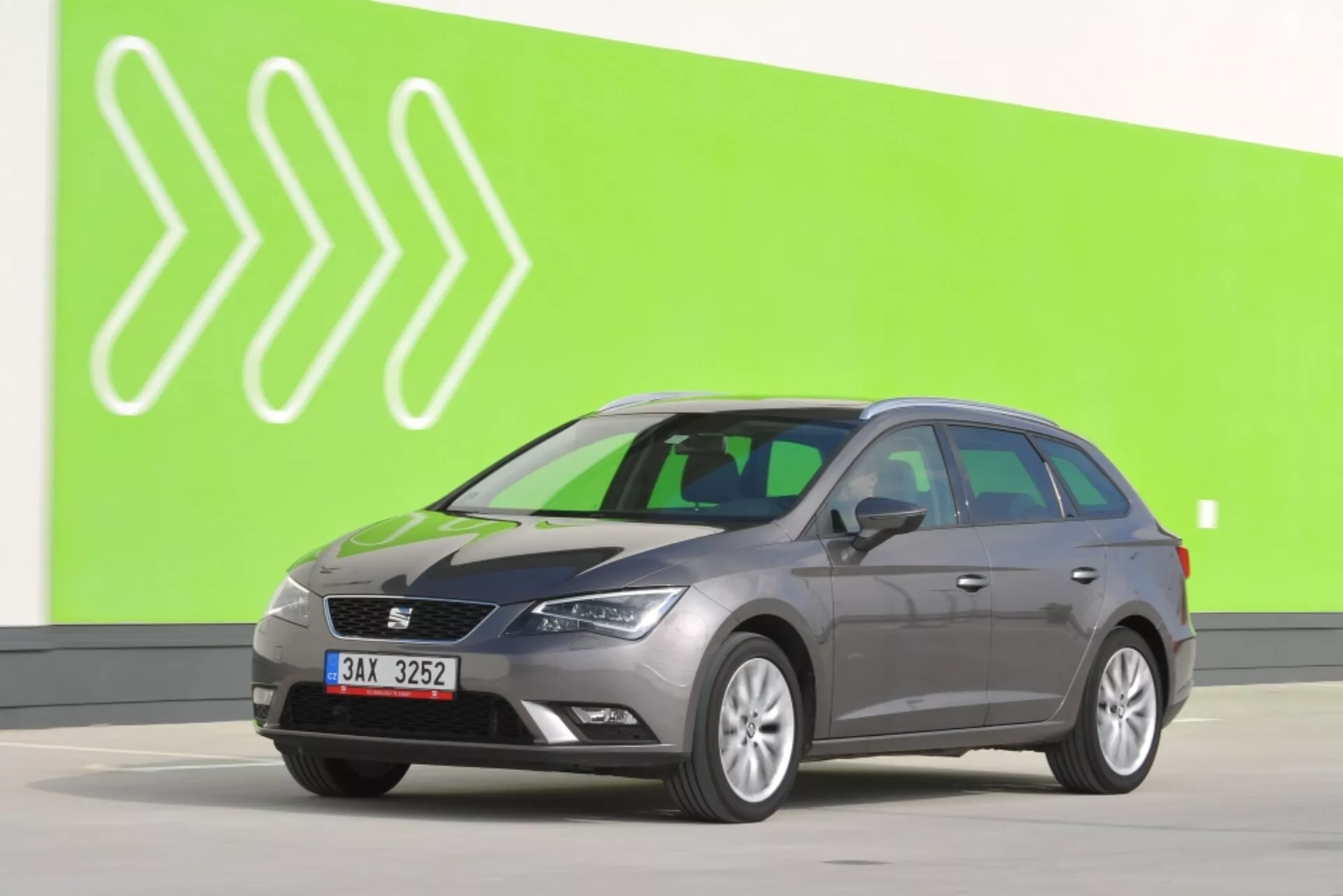 Szary kombi Seat Leon SC 2012 w pełnym widoku bocznym na zielonym tle ze stylizowanymi białymi groty strzał, prezentujący lusterka boczne z kierunkowskazami, charakterystyczne kontury drzwi i standardowe koła na jasnoszarej podłodze studia