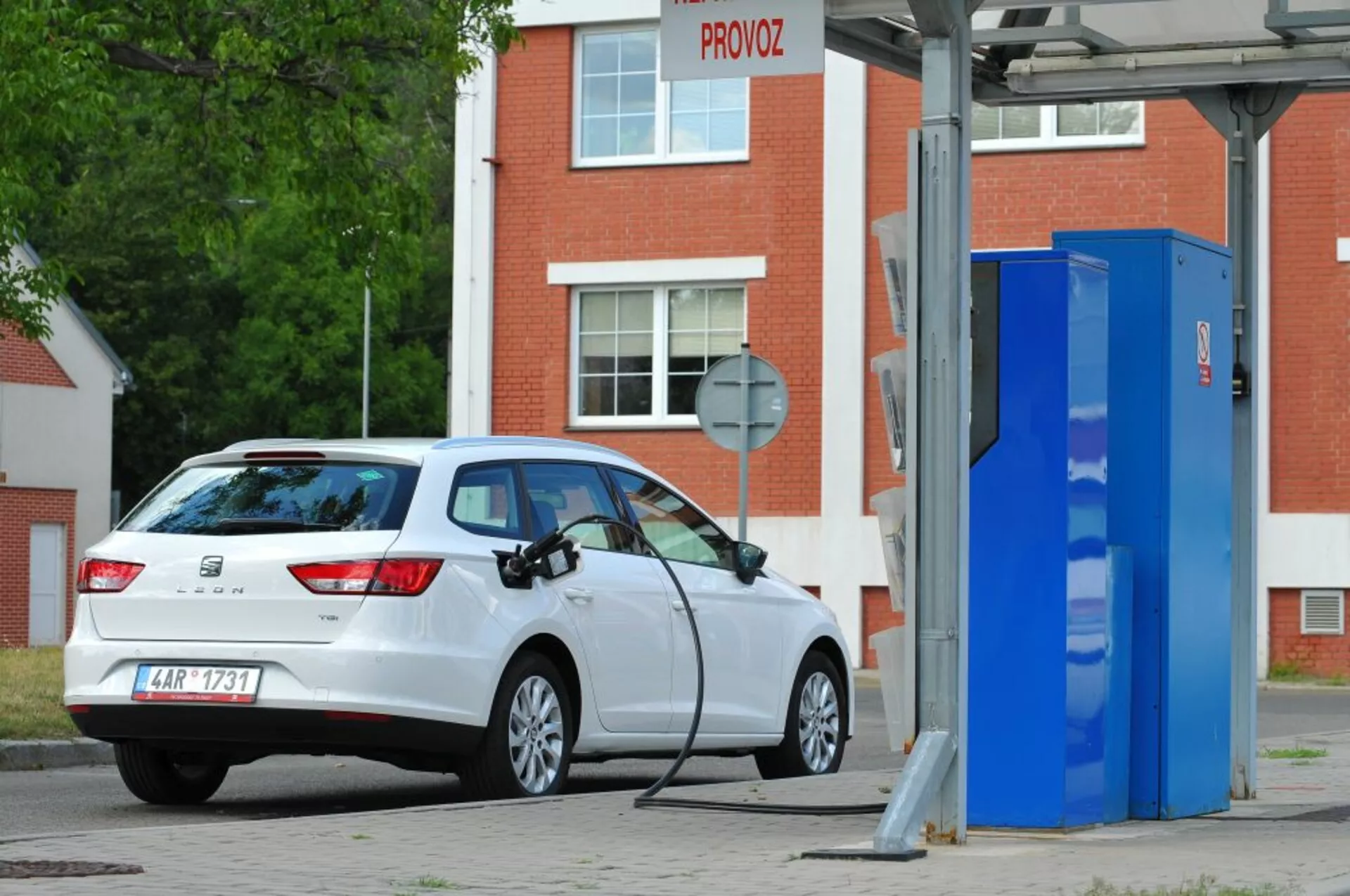 Biały hatchback Seat Leon SC z 2016 roku, elektryczny lub hybrydowy plug-in, zaparkowany przy stacji ładowania pojazdów elektrycznych, przedstawiający widok z tylnej trzeciej części podkreślający tylne światła, tylną szybę, profil klapy bagażnika i port ładowania po prawej stronie