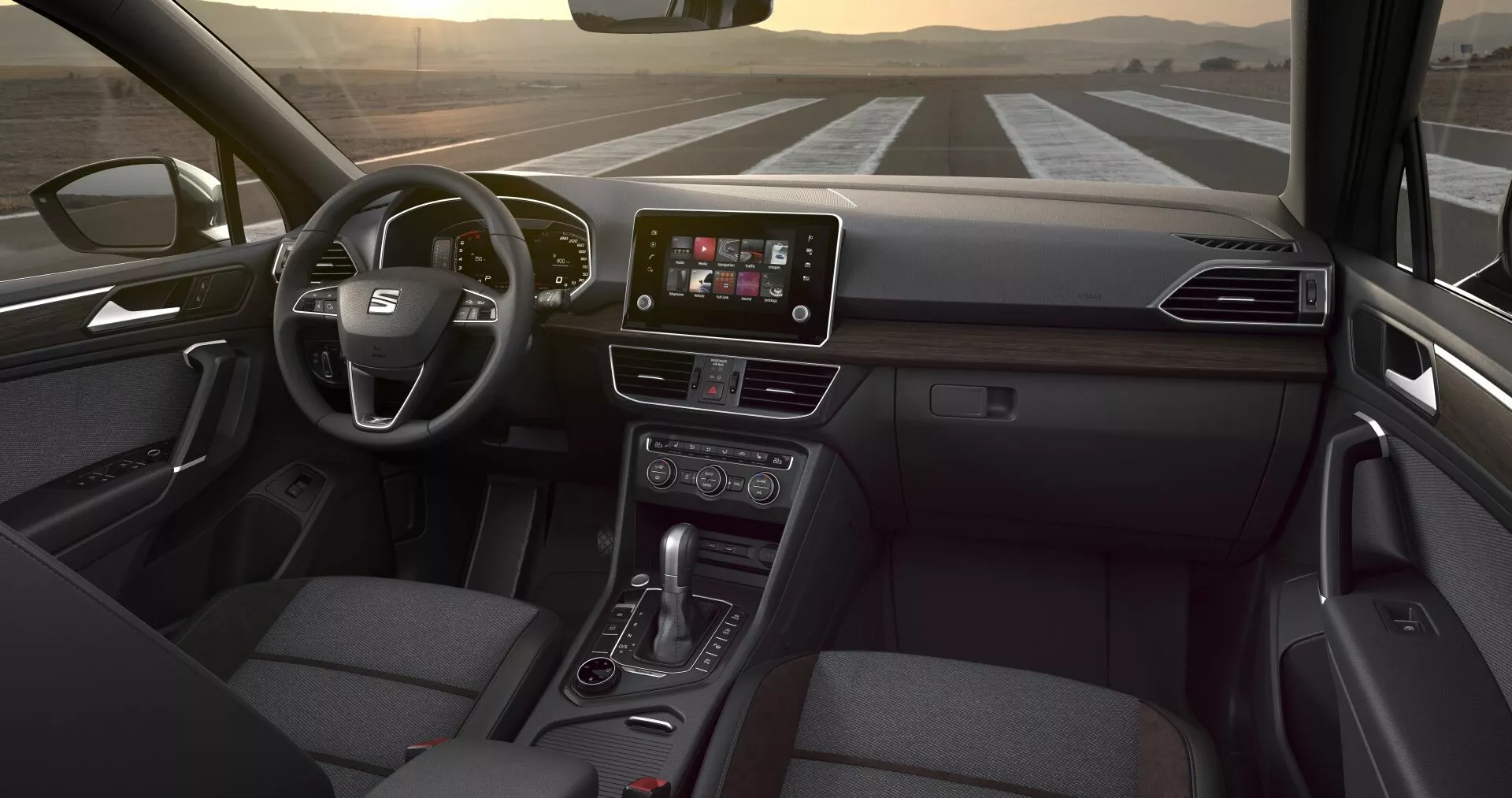 Perspektywa wnętrza Seat Tarraco 2019 z tylnego siedzenia, prezentująca wielofunkcyjną kierownicę, centralny ekran dotykowy systemu infotainment, przyciski sterowania klimatyzacją, dźwignię automatycznej skrzyni biegów i tapicerowane tkaniną siedzenie pasażera, przy subtelnych wewnętrznym oświetleniu