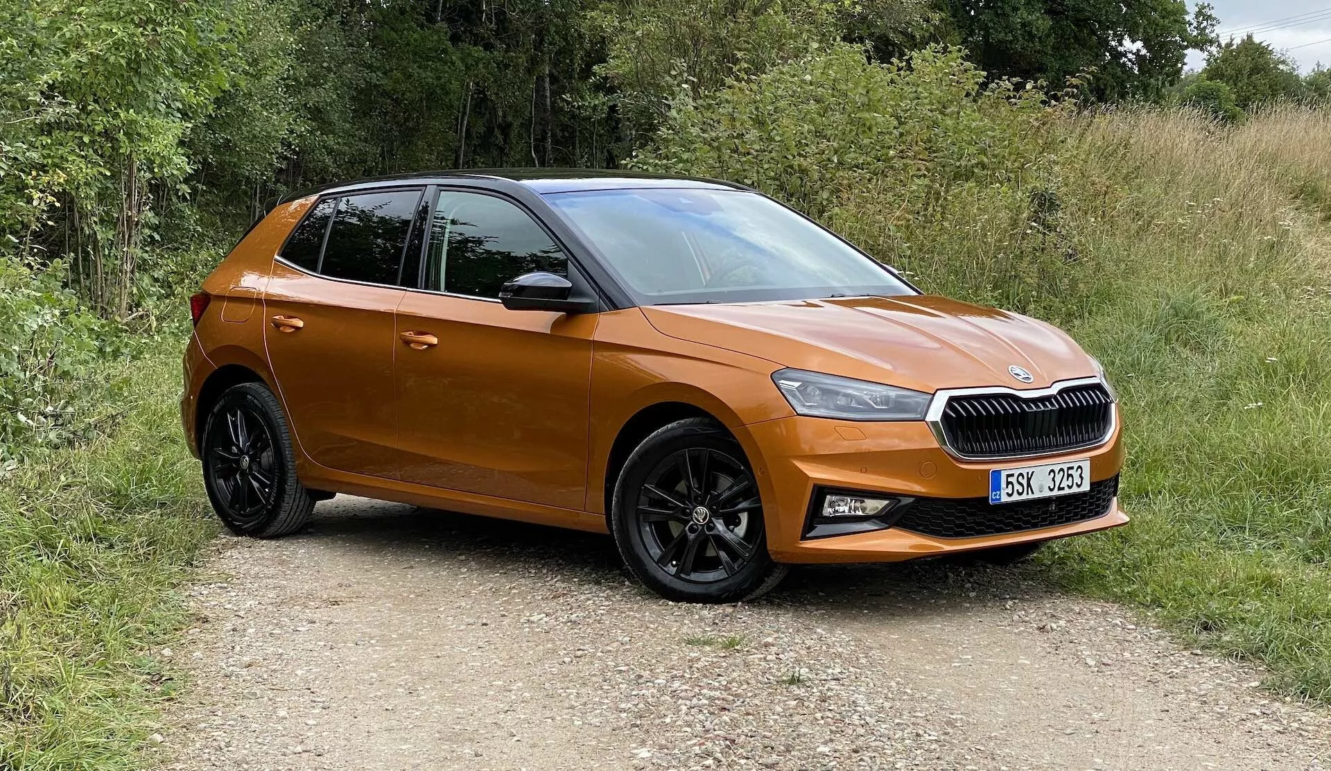 Zdjęcie nowoczesnego hatchbacka Škoda Fabia 2021, uchwycone z trzech czwartych przedniego widoku, ukazujące przedni lewy bok, drzwi kierowcy, lewą maskę i część dachu. Pojazd znajduje się na nieutwardzonej ścieżce otoczonej zielenią, co wskazuje na wiejskie otoczenie. Widoczne cechy to pozioma kratka z centralnym emblematem, ostre reflektory LED, metaliczny pomarańczowy lakier, lusterka boczne w kolorze nadwozia, czarne wieloszprychowe felgi z oponami z bieżnikiem oraz konturowane drzwi z aerodynamicznymi liniami. Samochód wygląda na nowy, kompaktowy i niestuningowany.
