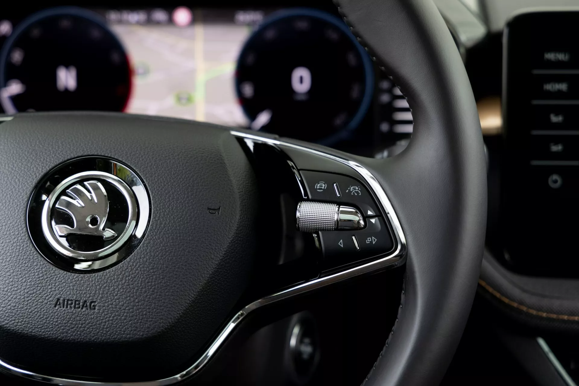 Zbliżenie wnętrza Skoda Fabia 2021 z perspektywy kierowcy, skupiając się na prawostronnych przyciskach kierownicy. Skórzana kierownica posiada centralny srebrny emblemat z logo Skoda i wytłoczony napis 'AIRBAG' poniżej. Zestaw przycisków po prawej stronie zawiera przyciski do obsługi systemu multimedialnego, połączeń bez użycia rąk lub komend głosowych, z manetkami widocznymi za kierownicą. Rozmyte tło przedstawia zestaw wskaźników z obrotomierzem i prędkościomierzem oraz centralnym wyświetlaczem cyfrowym. Konsola centralna lub system multimedialny częściowo widoczne po prawej stronie.
