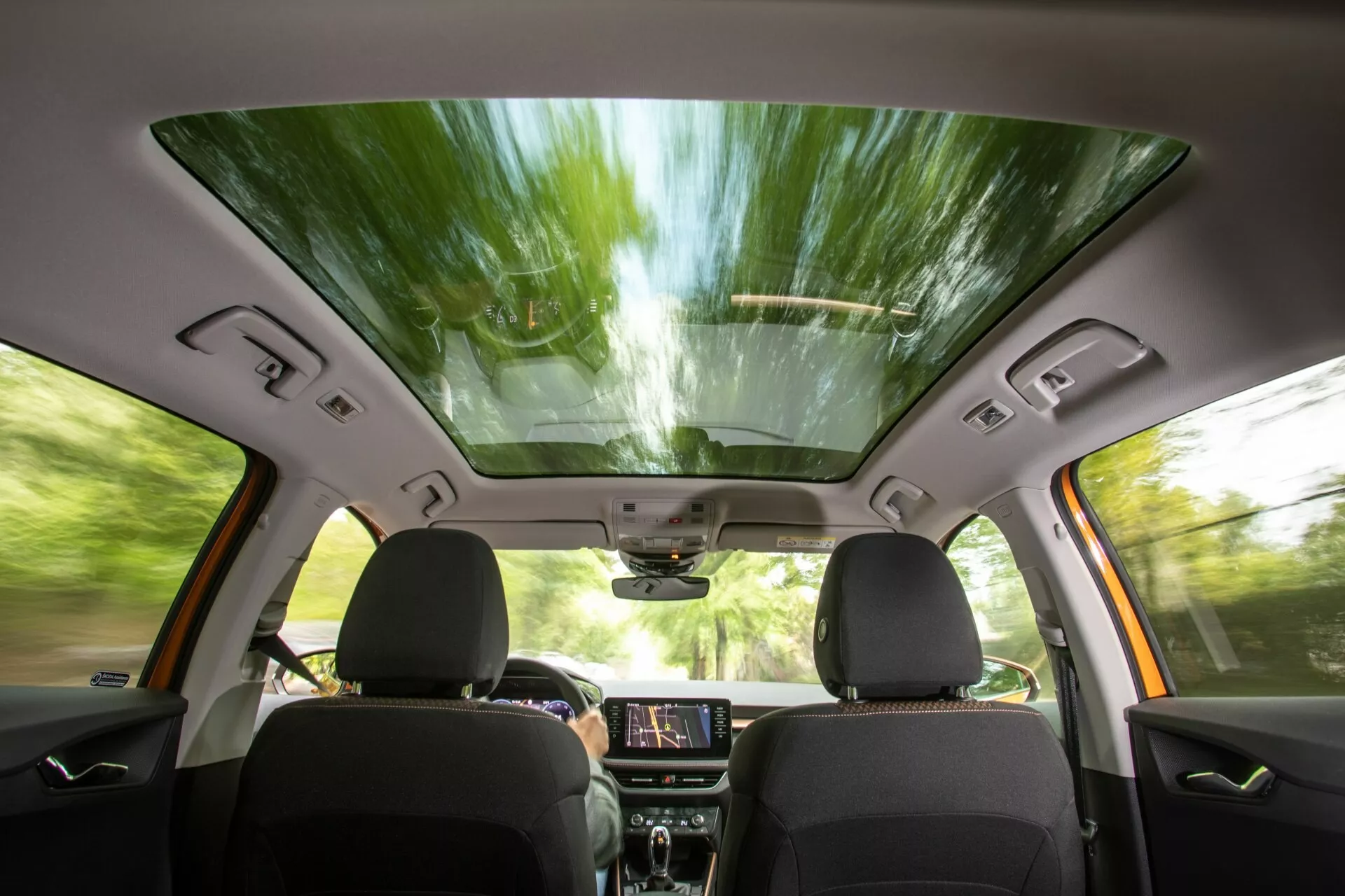 Widok wnętrza Skoda Fabia 2021 z perspektywy tylnego siedzenia, ukazujący przednie siedzenia z ciemną tapicerką, panoramiczny dach, podświetlony ekran dotykowy infotainmentu na desce rozdzielczej, panele sterowania, wbudowane w drzwi głośniki i sterowanie oknami, lusterko wsteczne z możliwymi funkcjami high-tech oraz konsolę górną do dodatkowych kontroli, sugerując nowoczesne, komfortowe i technologicznie zaawansowane doświadczenie jazdy.