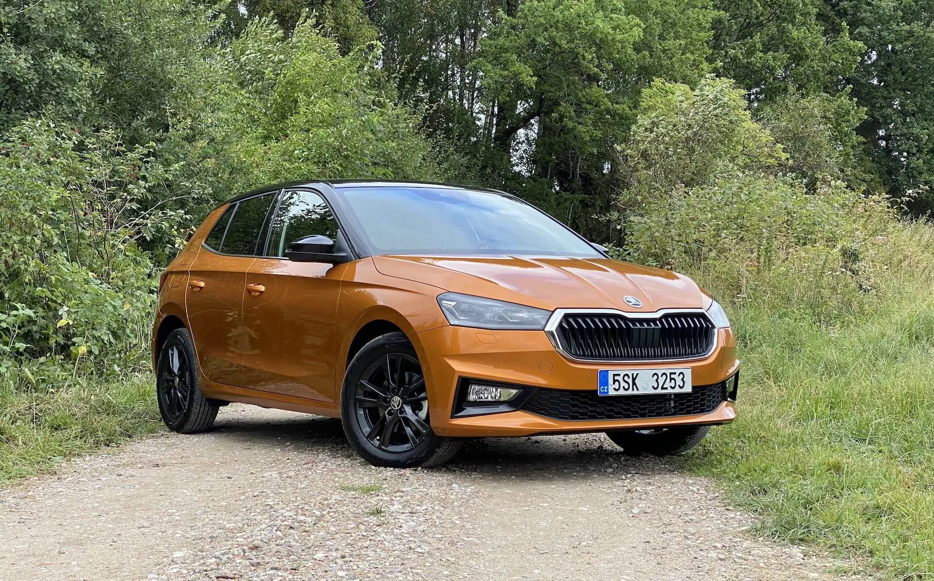 Pełen boczny profil nowoczesnego, pomarańczowego hatchbacka Skoda Fabia 2021 zaparkowanego na nieutwardzonej drodze z roślinnością w tle, uchwycony pod lekko skośnym kątem od frontu, podkreślając stronę kierowcy. Cechy obejmują wyraźny grill z centralnie umieszczonym emblematem marki, eleganckie reflektory, światła przeciwmgielne, duże czarne wieloramienne aluminiowe koła, klamki i lusterka w kolorze nadwozia, opadająca linia dachu i sugestię tylnej klapy oraz zespołu świateł.