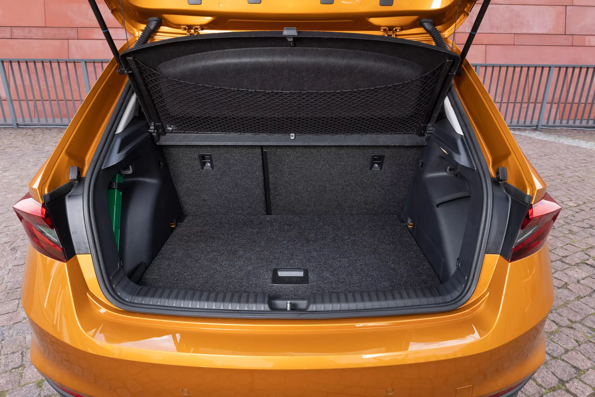 Obraz przedstawia bagażnik samochodu Skoda Fabia model 2021, uchwycony z lekko podwyższonego tylnego kąta, aby pokazać przestronną przestrzeń ładunkową. Wnętrze jest wyłożone materiałem przypominającym dywan i wyposażone w elastyczne boczne miejsca do przechowywania, przegrodę z siatki na ładunek oraz wnękę w dolnej części na dodatkowe przedmioty. Klapa bagażnika jest podniesiona, ukazując duży czysty bagażnik, zaprojektowany aby podkreślić pojemność bagażową i praktyczność pojazdu.