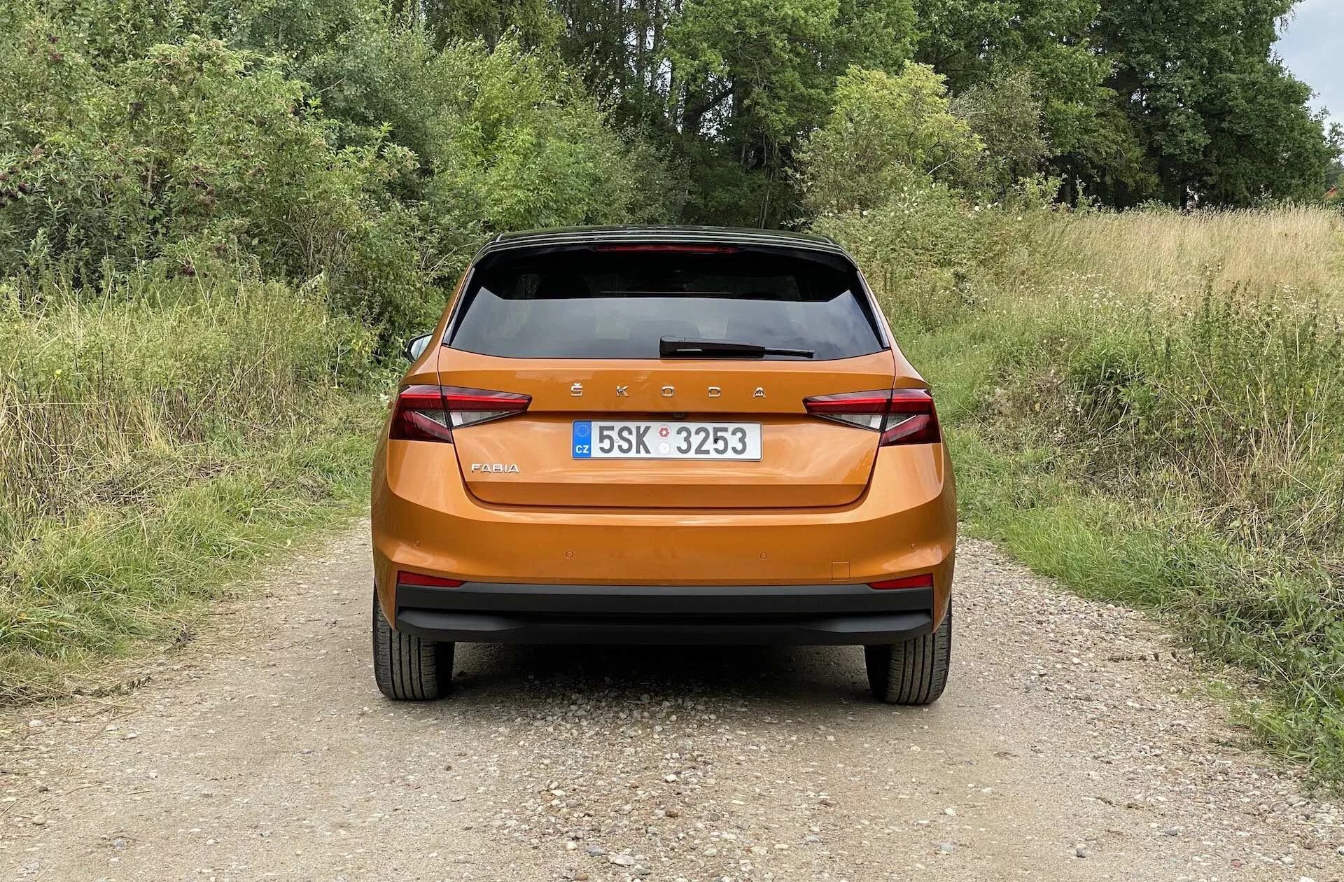 Widok z tyłu jasnopomarańczowego metalicznego hatchbacka Skoda Fabia 2021, sfotografowanego w świetle dziennym na wąskiej, gruntowej ścieżce w wiejskiej scenerii. Zdjęcie uchwyca nowoczesny tylny design z ciągłym tylnym pasem świetlnym, ostrymi kanciastymi światłami tylnymi, zintegrowanymi odblaskami, tylną wycieraczką i tylnym spojlerem poprawiającym aerodynamikę, podkreślającym błyszczące wykończenie i praktyczne funkcje samochodu.