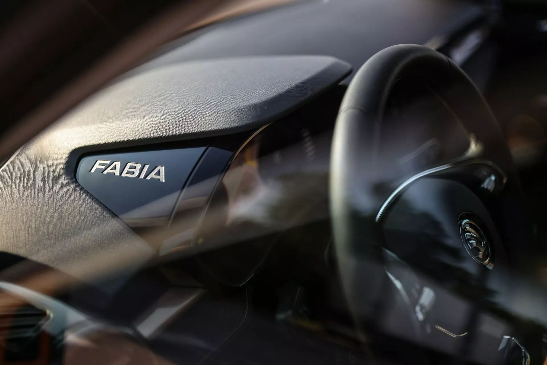 Zbliżenie wnętrza Skody Fabia 2021, z uwydatnioną prawą stroną deski rozdzielczej. Wyraźnie widoczny jest napis 'FABIA', podczas gdy nowoczesna kierownica z logo Skoda jest częściowo widoczna po lewej stronie. Deska rozdzielcza ma ziarnistą teksturę, a płytka głębia ostrości podkreśla znak fabryczny na lekko rozmytym tle.