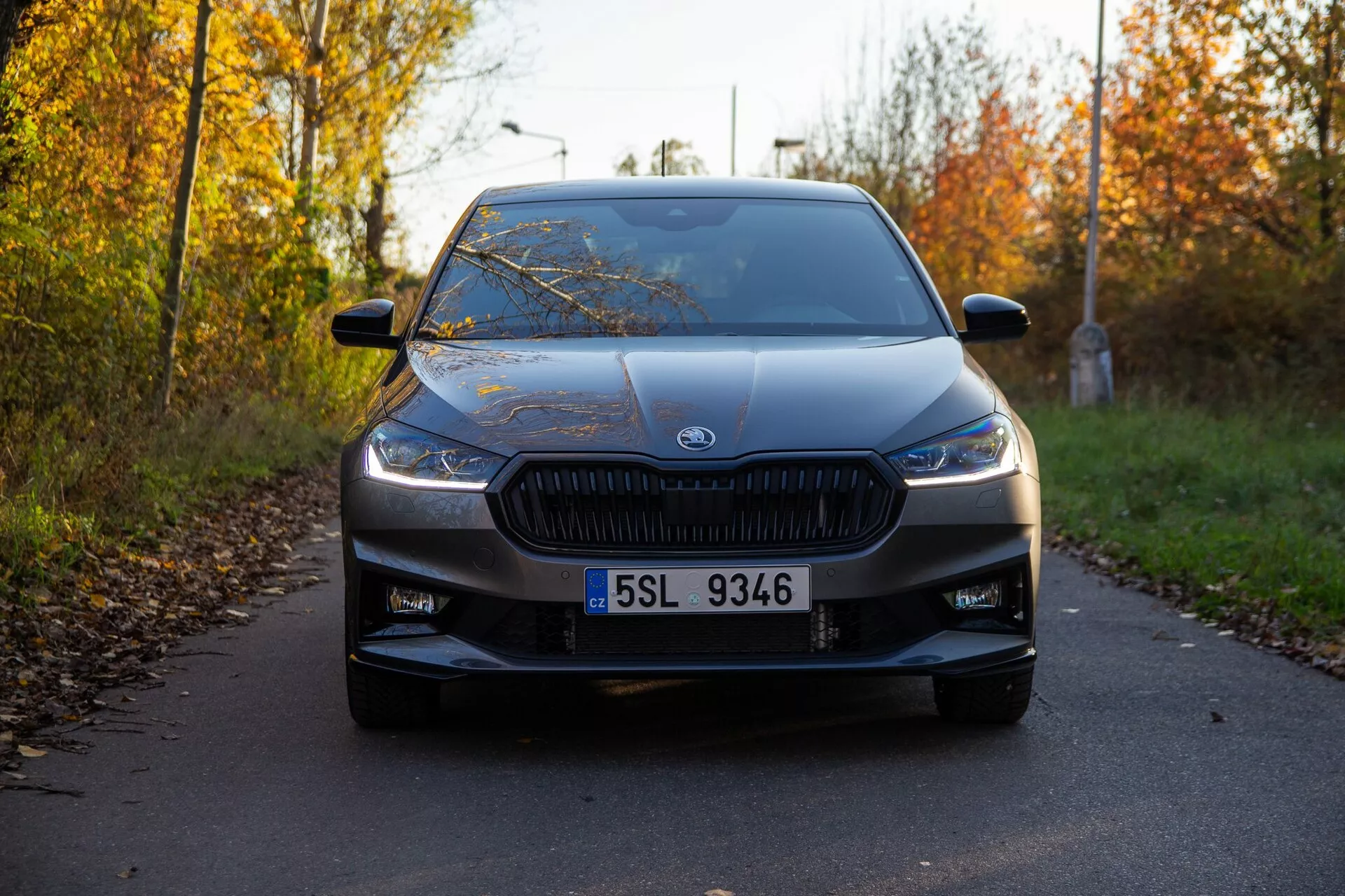 Widok z przodu Skoda Fabia 2021 uchwycony z niskiego kąta, podkreślający charakterystyczną kratkę z pionowymi listwami i nowoczesnymi, wąskimi reflektorami. Samochód, ustawiony na środku kadru, jest zaparkowany na wąskiej drodze otoczonej jesiennym listowiem, z wyraźnie widocznym emblematem producenta umieszczonym na środku kratki. Dodatkowe widoczne szczegóły projektowe obejmują czytelną tablicę rejestracyjną, wloty powietrza w dolnym zderzaku, delikatne linie konturowe na masce, rozległą przednią szybę i lusterka boczne z kierunkowskazami. Miękkie światło i długie cienie sugerują, że zdjęcie zostało zrobione wcześnie rano lub późnym popołudniem, ukazując samochód w doskonałym stanie.