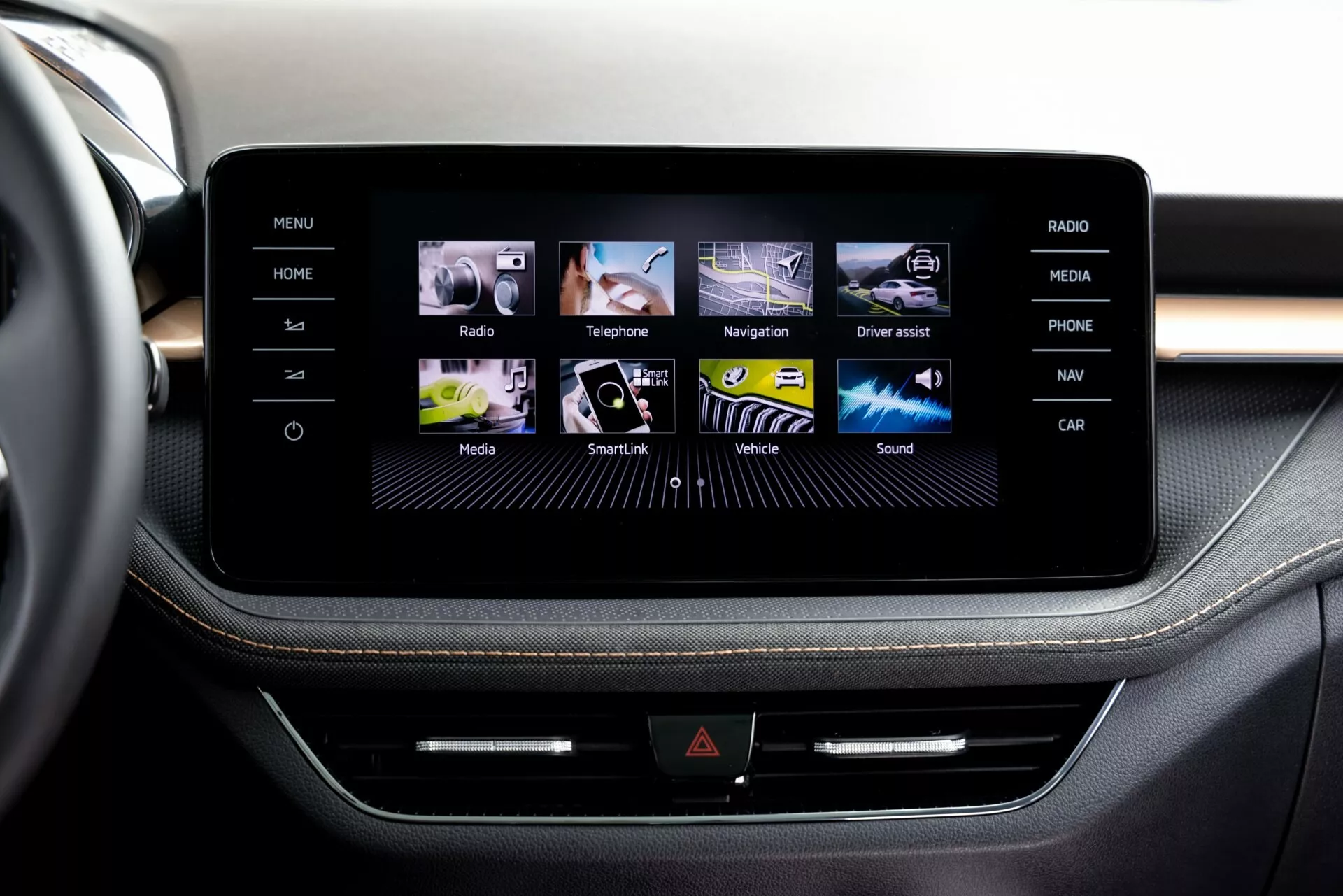 Zbliżenie na system infotainment z ekranem dotykowym w modelu Škoda Fabia 2021 z perspektywy pasażera lub kierowcy siedzącego w pierwszym rzędzie, ukazujący elegancką konsolę środkową obramowaną przypuszczalnie szczotkowanym aluminium. Ekran wyświetla przyjazny dla użytkownika interfejs graficzny z kolorowymi ikonami dla funkcji takich jak Dom, Radio, Telefon, Nawigacja i inne. Poniżej ekranu znajduje się linia przycisków fizycznych i możliwe, że gniazdo CD, które dodaje wielofunkcyjności. Widoczna tapicerka o wykończeniu premium z kontrastującymi szwami wskazuje na wysoki standard wnętrza pojazdu.