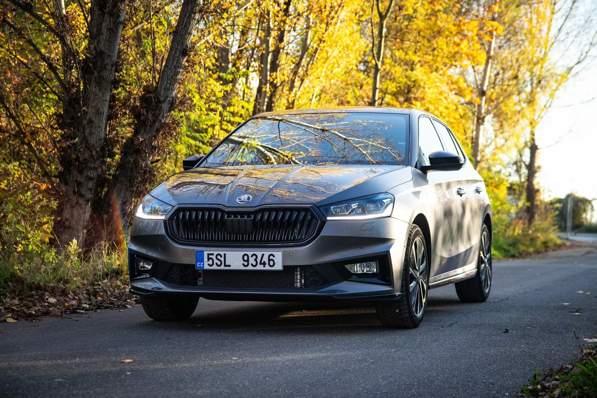 Obraz nowoczesnego sedana Skoda Fabia 2021 zaparkowanego na drodze otoczonej jesiennym listowiem, uchwycony z perspektywy na poziomie gruntu, podkreślający przednią kratkę, kątowe reflektory, eleganckie linie projektowe, aluminiowe felgi i boczne lusterka z wbudowanymi kierunkowskazami, odzwierciedlające wysoką jakość wykończenia metalicznego oraz połączenie luksusu i sportowego charakteru.