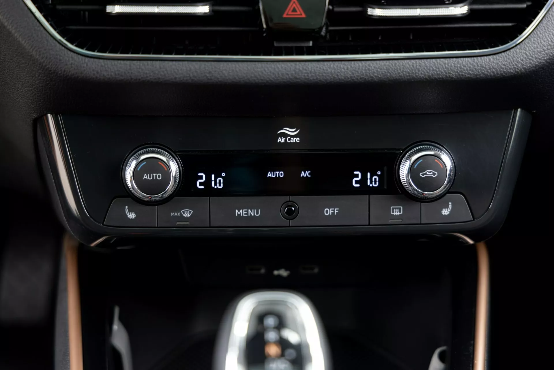 Zbliżenie na panel klimatyzacji Skoda Fabia 2021 umieszczony na konsoli środkowej, z dwustrefowymi kontrolerami temperatury z wyświetlaczami cyfrowymi pokazującymi '20', zintegrowanymi przyciskami do automatycznej kontroli klimatu ('AUTO'), klimatyzacji ('AC'), ustawieniami menu ('MENU') i wyłączaniem systemu ('OFF'). Błyszcząca czarna ramka otacza wyświetlacz informacyjny nad kontrolkami, z widocznym przyciskiem ostrzegawczym (ikona trójkąta) na górze. Panel jest umieszczony w wysokiej jakości wnętrzu z matowym wykończeniem i elementami przypominającymi drewno, podkreślając nowoczesny design i technologię.