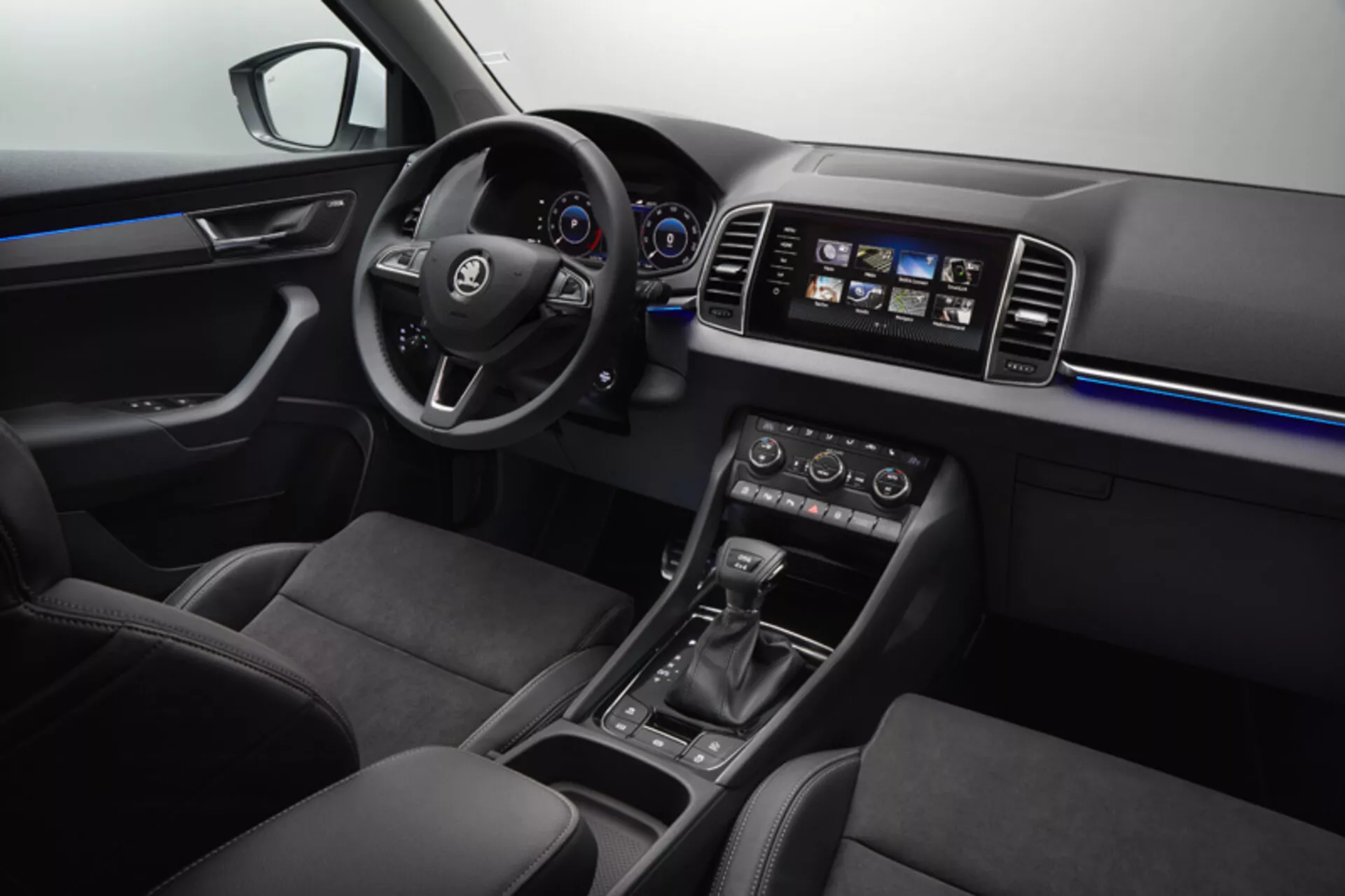 Widok wnętrza Skoda Karoq 2017 przedstawiający kierownicę z zamontowanymi sterowaniami, cyfrowy zegar, cechy konsoli środkowej, system infotainment dotykowy, klimatyzację dwustrefową, ambientowe oświetlenie wnętrza, sportowe przednie siedzenia oraz eleganckie wykończenie łączące miękkie tworzywa, akcenty z szczotkowanego metalu i panele wysokiego połysku