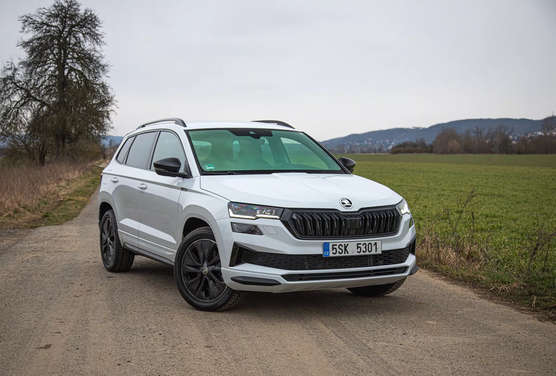 Biały SUV Škoda Karoq 2021 zaparkowany na wiejskiej drodze gruntowej, prezentujący widok z przedniej lewej ćwiartki włącznie z grillem, reflektorami i projektowaniem kół, ze szczególnym uwzględnieniem nowoczesnych funkcji, takich jak oświetlenie LED i czujniki parkowania.