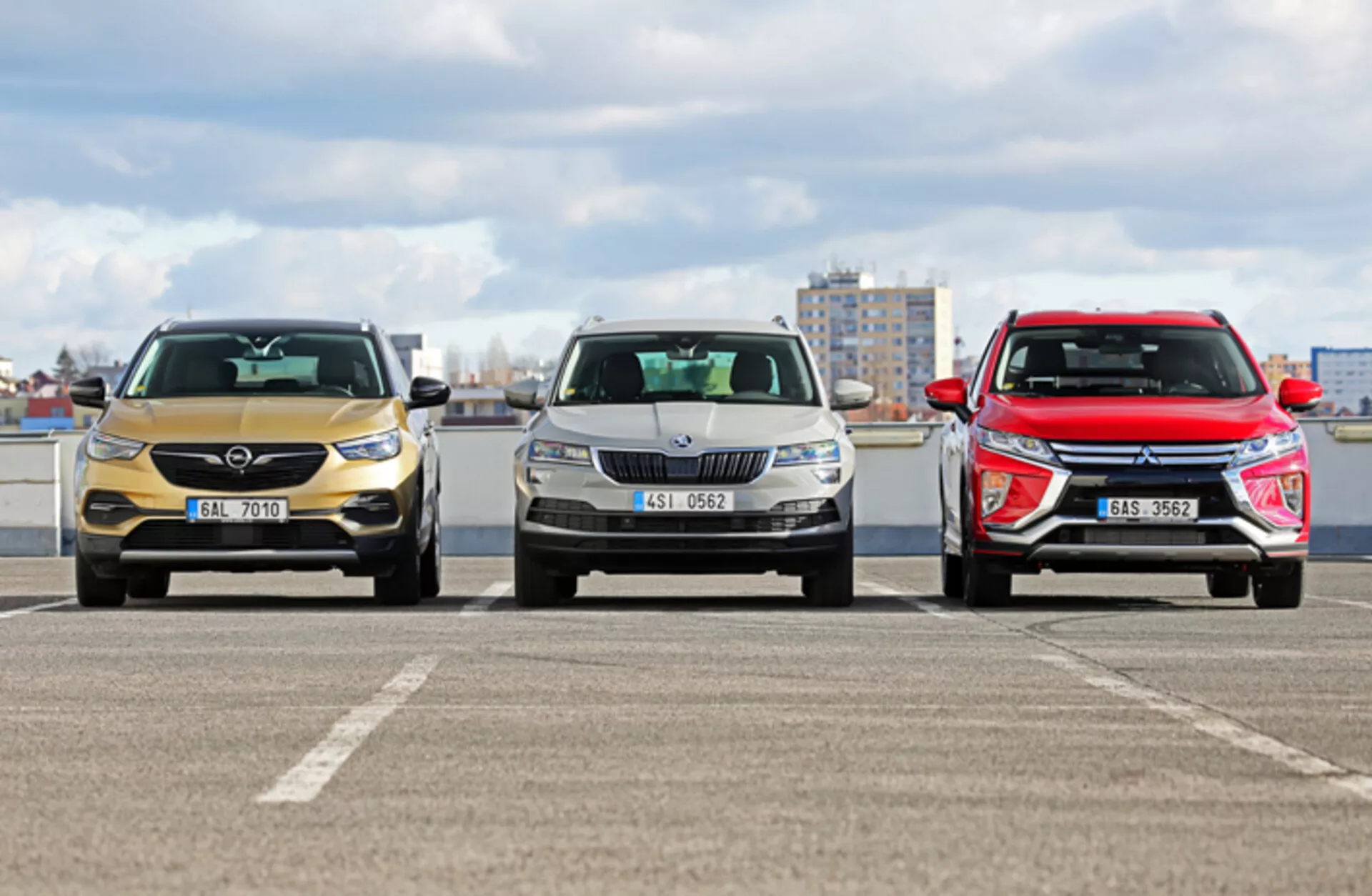 Przedni widok trzech modeli Skoda Karoq 2017 na parkingu z tłem krajobrazu miejskiego, prezentujący różne wzory grilla, reflektorów i kształty aerodynamiczne maski w warunkach pochmurnego światła dziennego