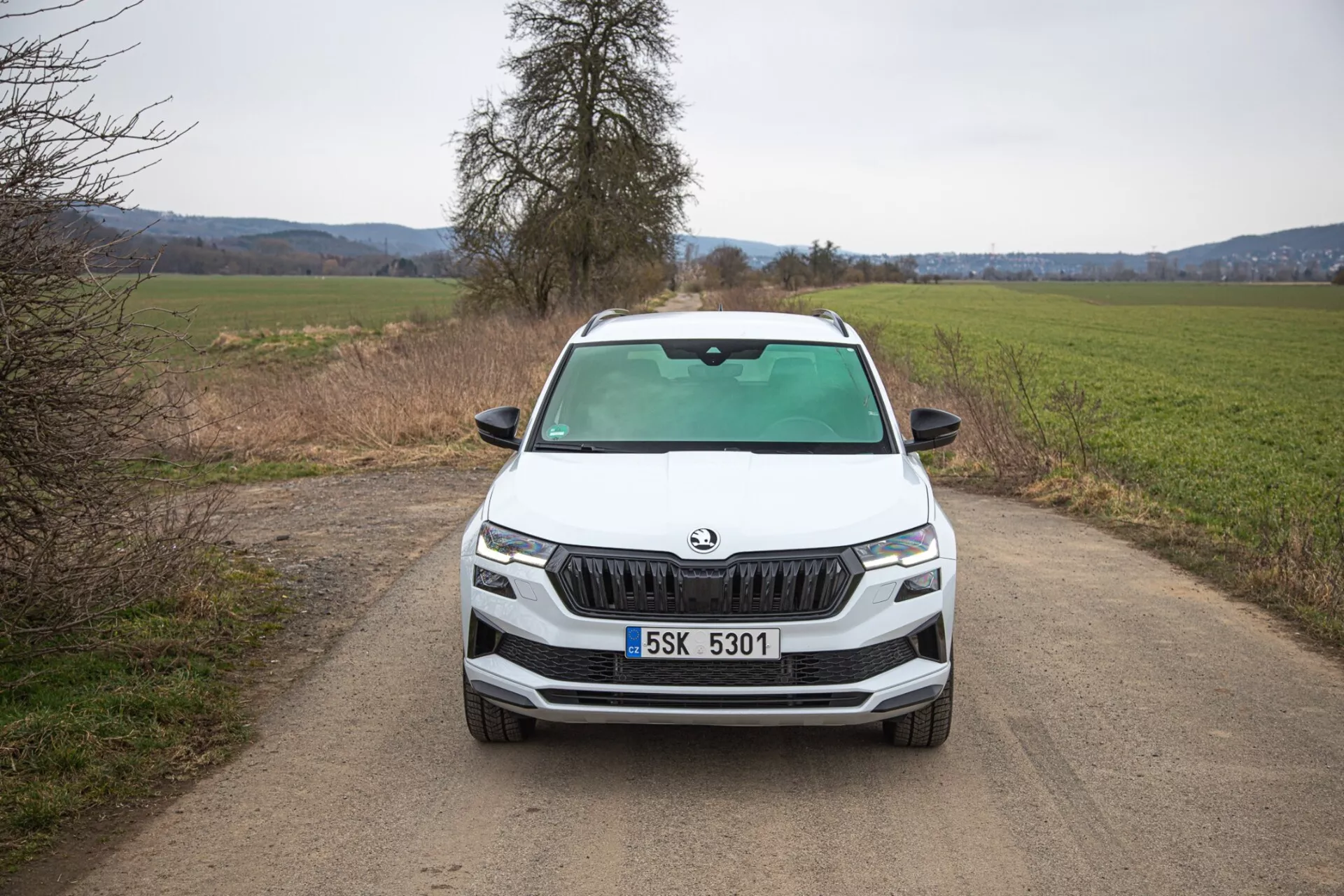 SUV Škoda Karoq 2021 przedstawiający bezpośredni widok frontalny na wąskiej, nieutwardzonej drodze wiejskiej, podkreślający charakterystyczną atrapę Škody, ostre reflektory LED, światła przeciwmgielne w czarnej obwódce sugerujące potencjalne zdolności terenowe, dynamiczne linie maski oraz zaawansowane funkcje technologiczne na przednim zderzaku.