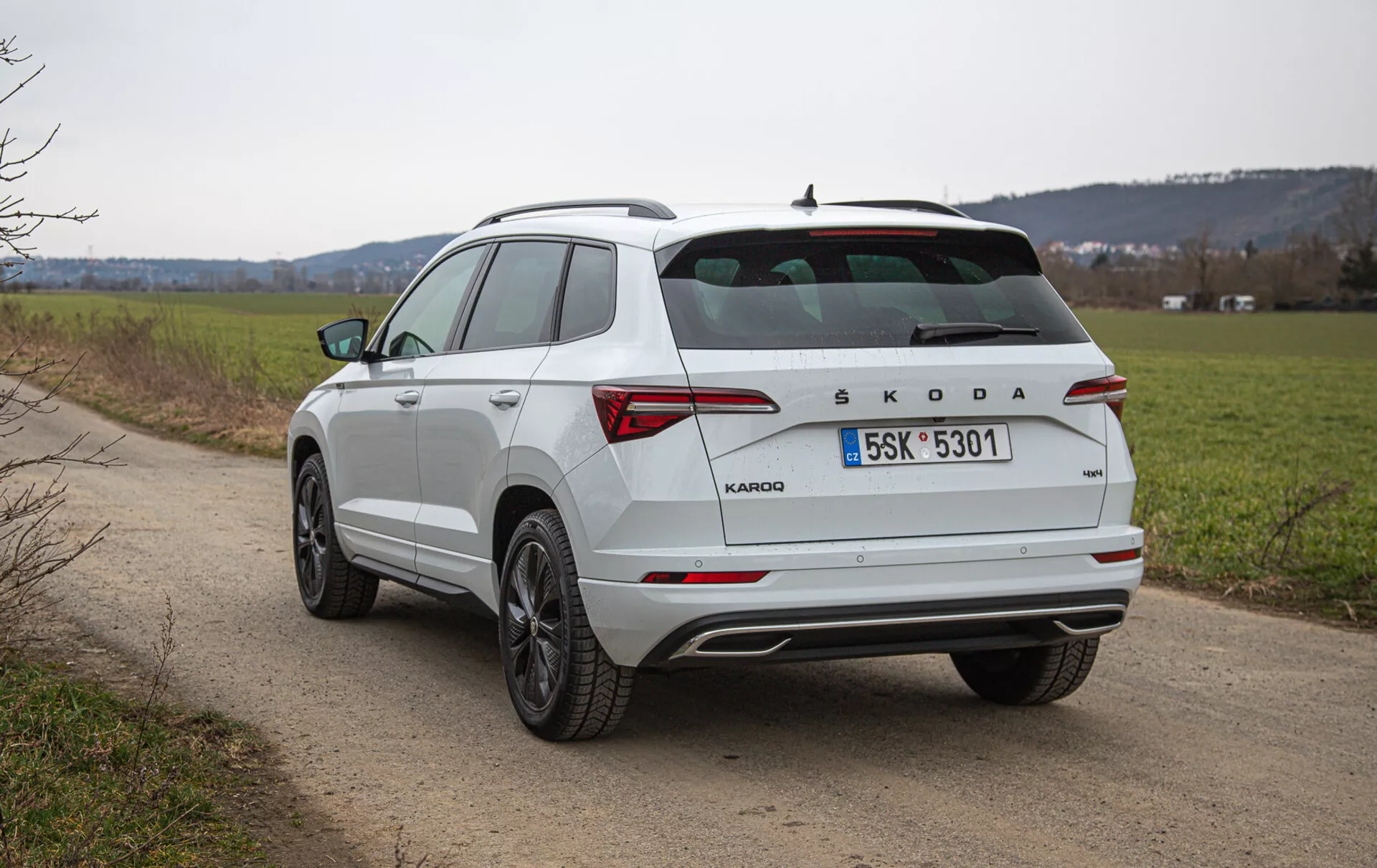 Widok z tyłu z trzech czwartych na biały, kompaktowy SUV Škoda Karoq na wiejskiej drodze, z nachyloną tylną szybą, relingami dachowymi, tylnymi światłami LED, brandingiem Škoda, alufelgami wieloramiennymi i chromowanymi detalami, sfotografowany na zewnątrz w świetle dziennym z naturalnym krajobrazem w tle.