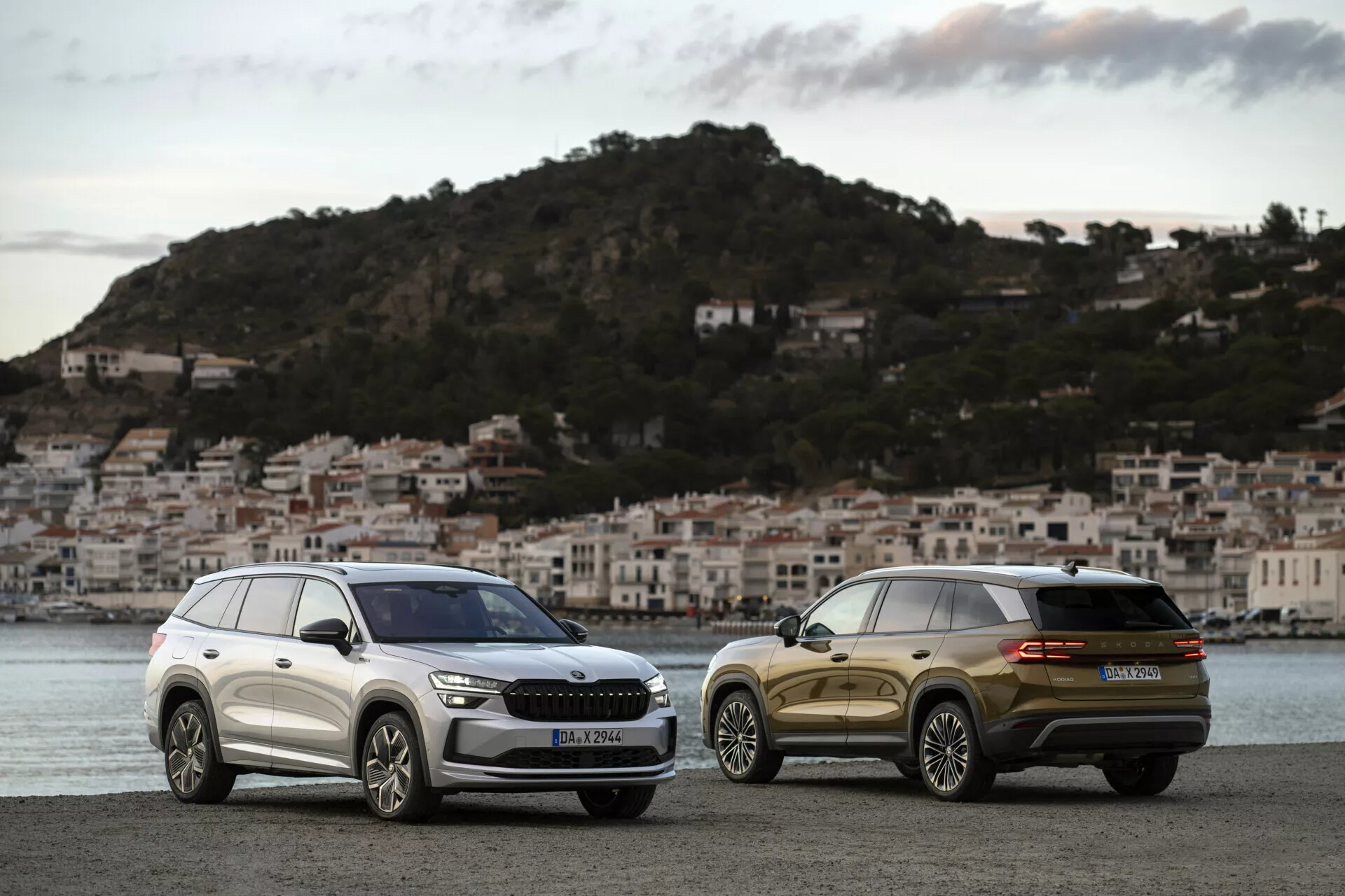 SUVy Skoda Kodiaq 2024 zaparkowane obok siebie o świcie na promenadzie nadmorskiej z miasteczkiem nadmorskim w tle, prezentujące grill, reflektory i eleganckie linie designu jednego oraz widok z tyłu trzy czwarte drugiego.