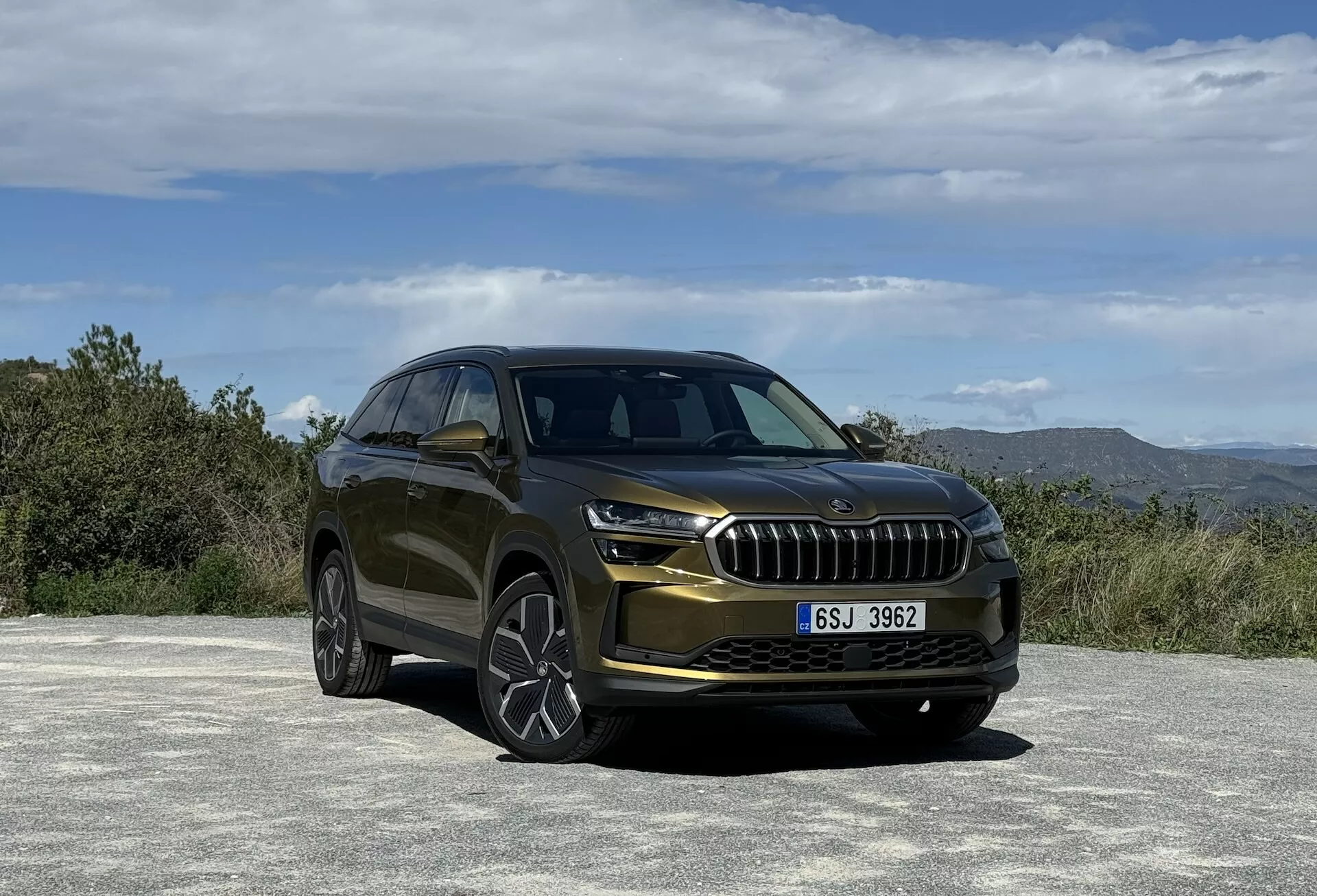 SUV Skoda Kodiaq 2024 z widoku trzy czwarte z przodu podkreślający jego luksusową kratę, eleganckie reflektory LED, sportowe obudowy świateł przeciwmgielnych i wloty powietrza, umieszczony na tle krajobrazu naturalnego z metalicznym wykończeniem lakieru i felgami aluminiowymi o wielu szprychach.